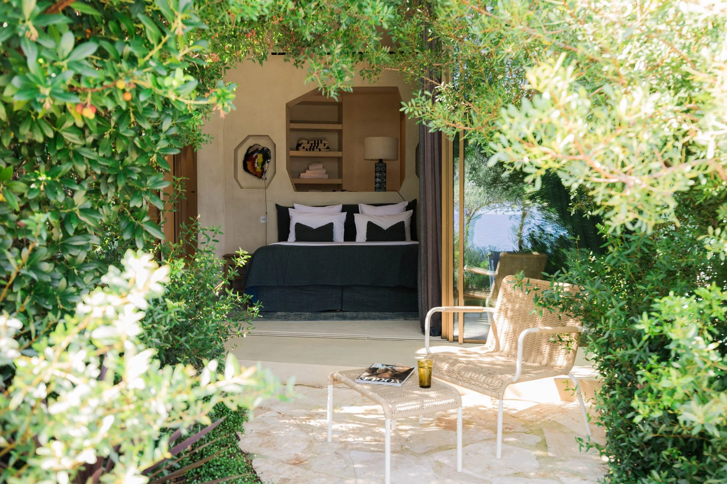 A cozy bedroom with a black bed, white pillows, and a view of an outdoor patio with two woven chairs, a small table with a magazine and a drink, surrounded by greenery.