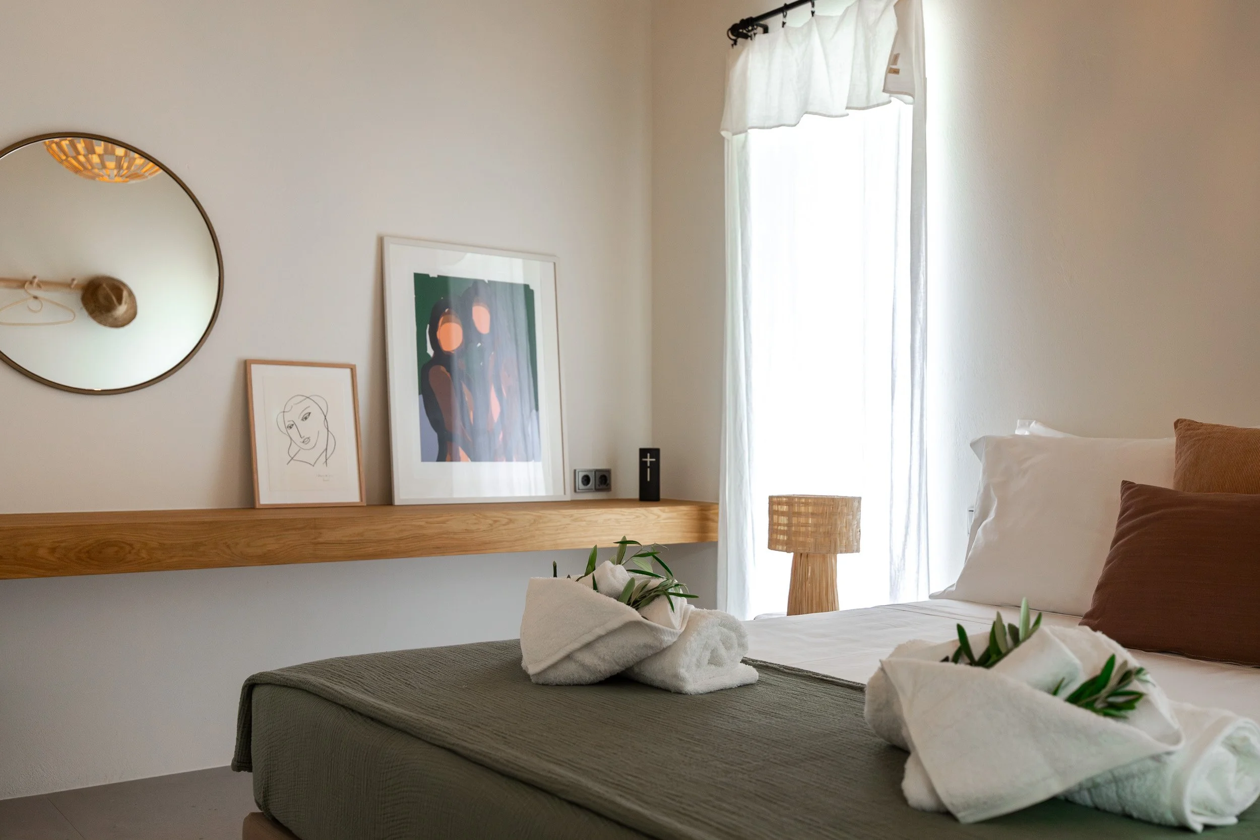 A neatly made bed with white linens, brown pillows, and towel art with greenery on top. A window with sheer white curtains allows natural light into the room. A wooden nightstand with a woven lamp sits beside the bed. There is a wooden ledge on the w