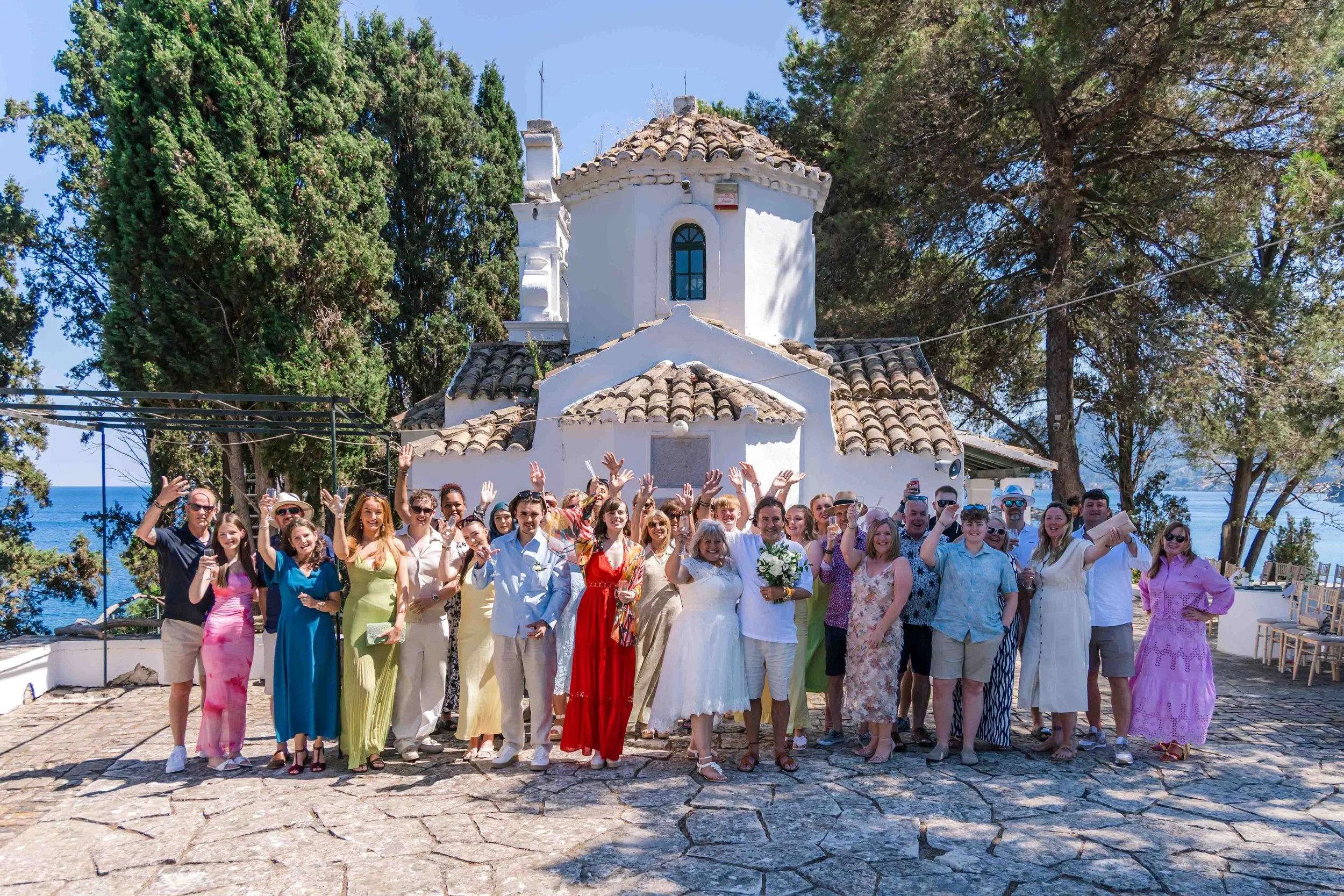 A large group of people celebrating a wedding outside a small white chapel with a tiled roof near the sea, with trees and blue sky in the background.