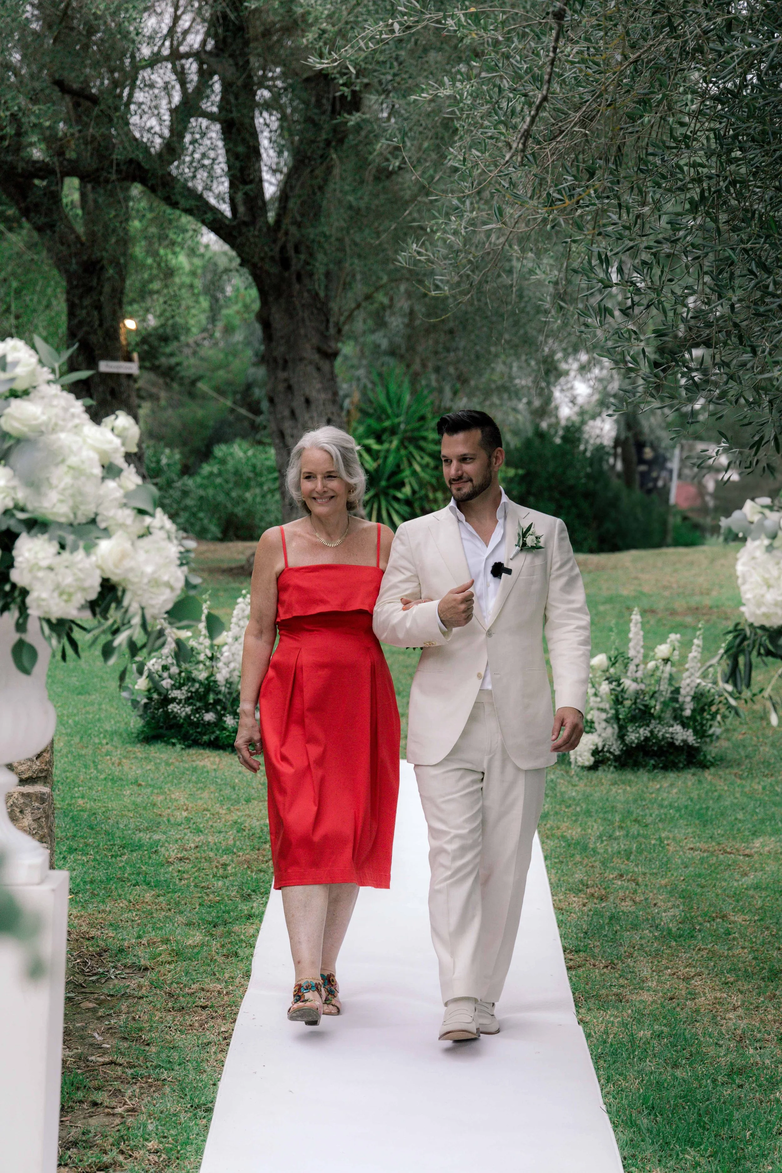A man in a white suit walking down an outdoor aisle with an older woman in a red dress, surrounded by white flowers and green trees.