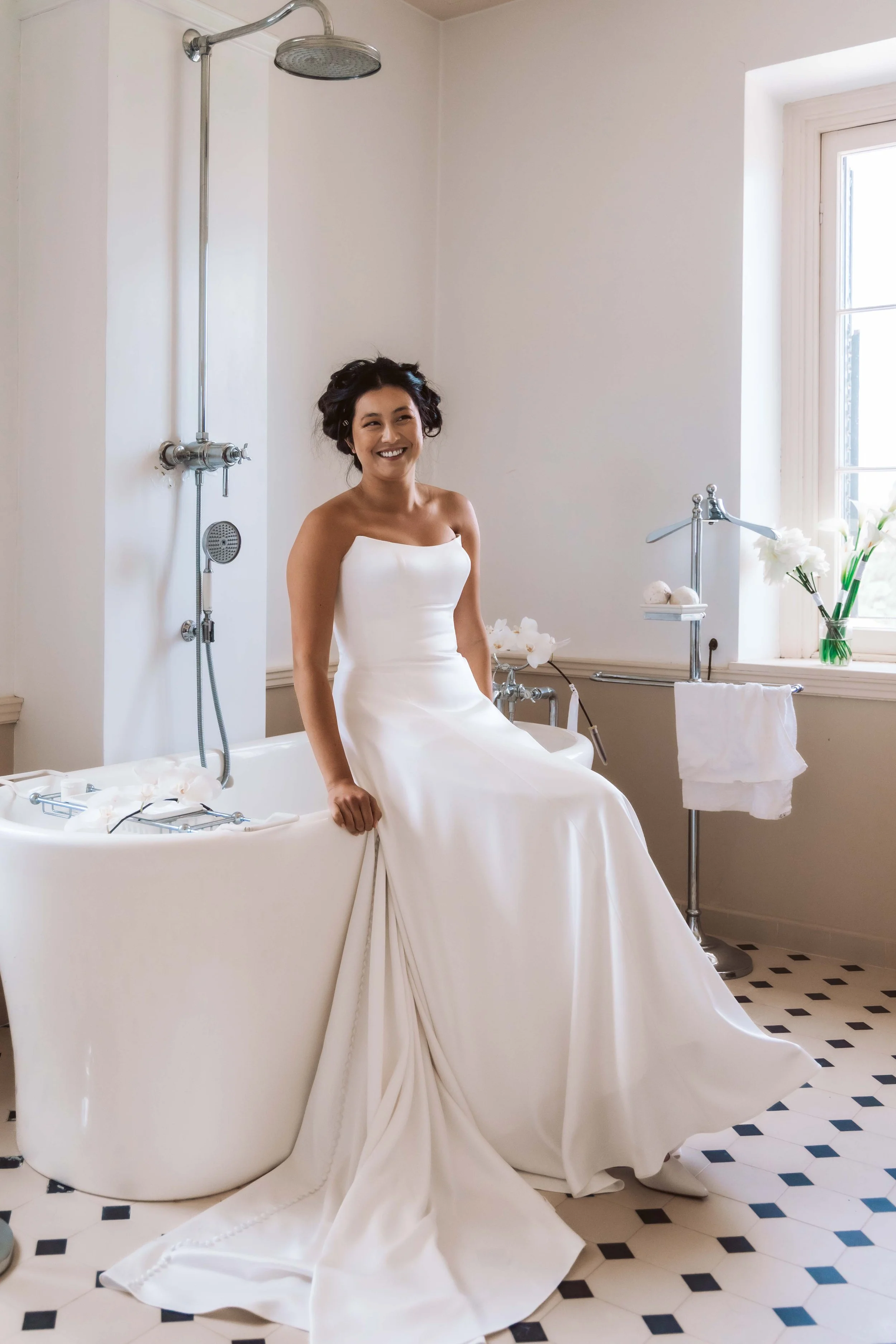 A woman in a white wedding dress sitting on the edge of a bathtub in a bright, minimalist bathroom with a window, flowers, and bathroom accessories.