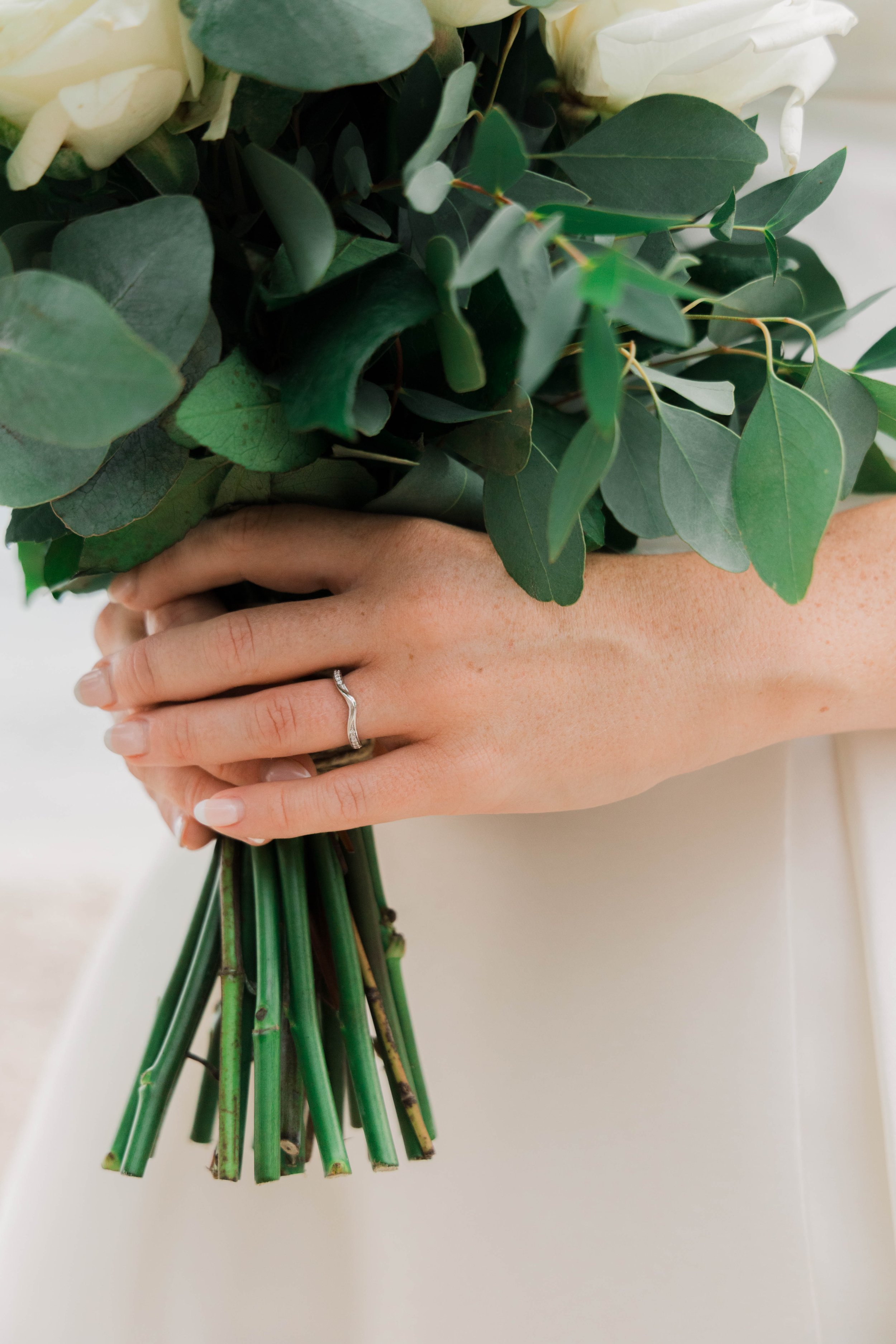 Close-up of a woman's hand holding a bouquet of green leaves and white flowers, showing a silver ring on her ring finger.