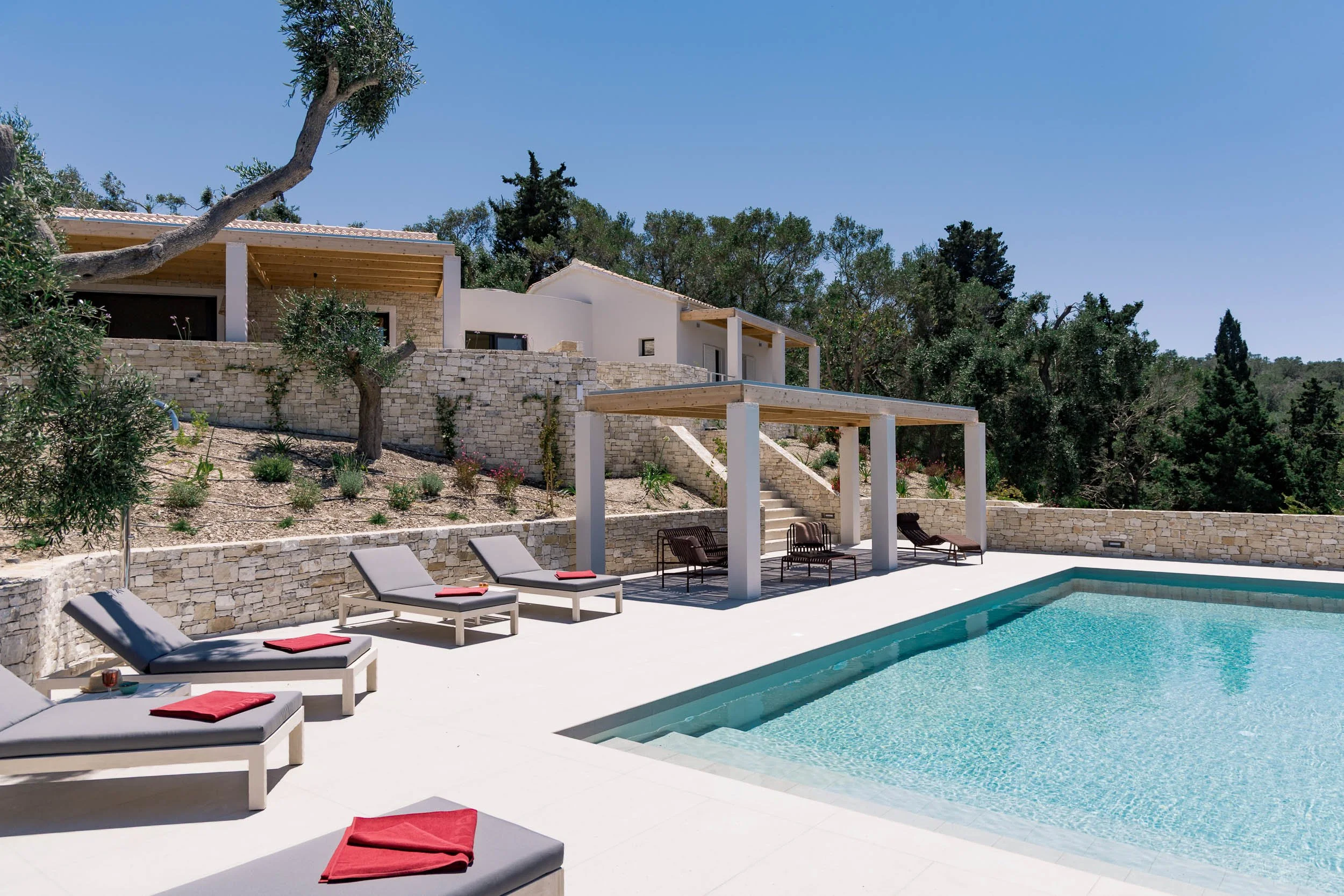 Empty outdoor swimming pool area with lounge chairs, some with red towels, under a shaded structure. The background features a modern white house with stone walls, stairs, and lush green trees under a clear blue sky.