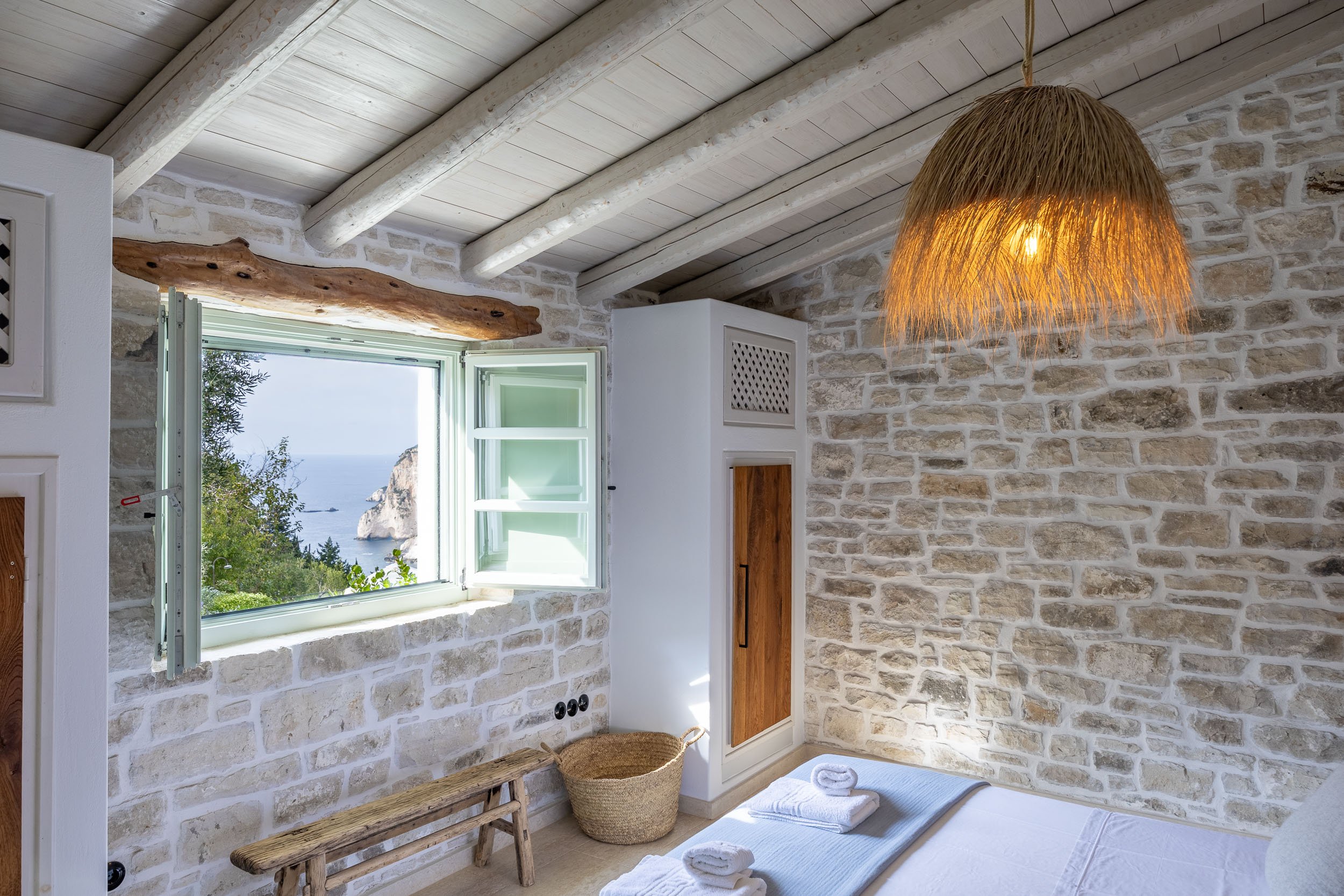 A cozy bedroom with white brick walls, a wooden ceiling with exposed beams, a window showing a scenic view of the ocean and cliffs, a woven hanging light, a small white wooden cabinet, a woven basket on the floor, and folded towels on the bed.