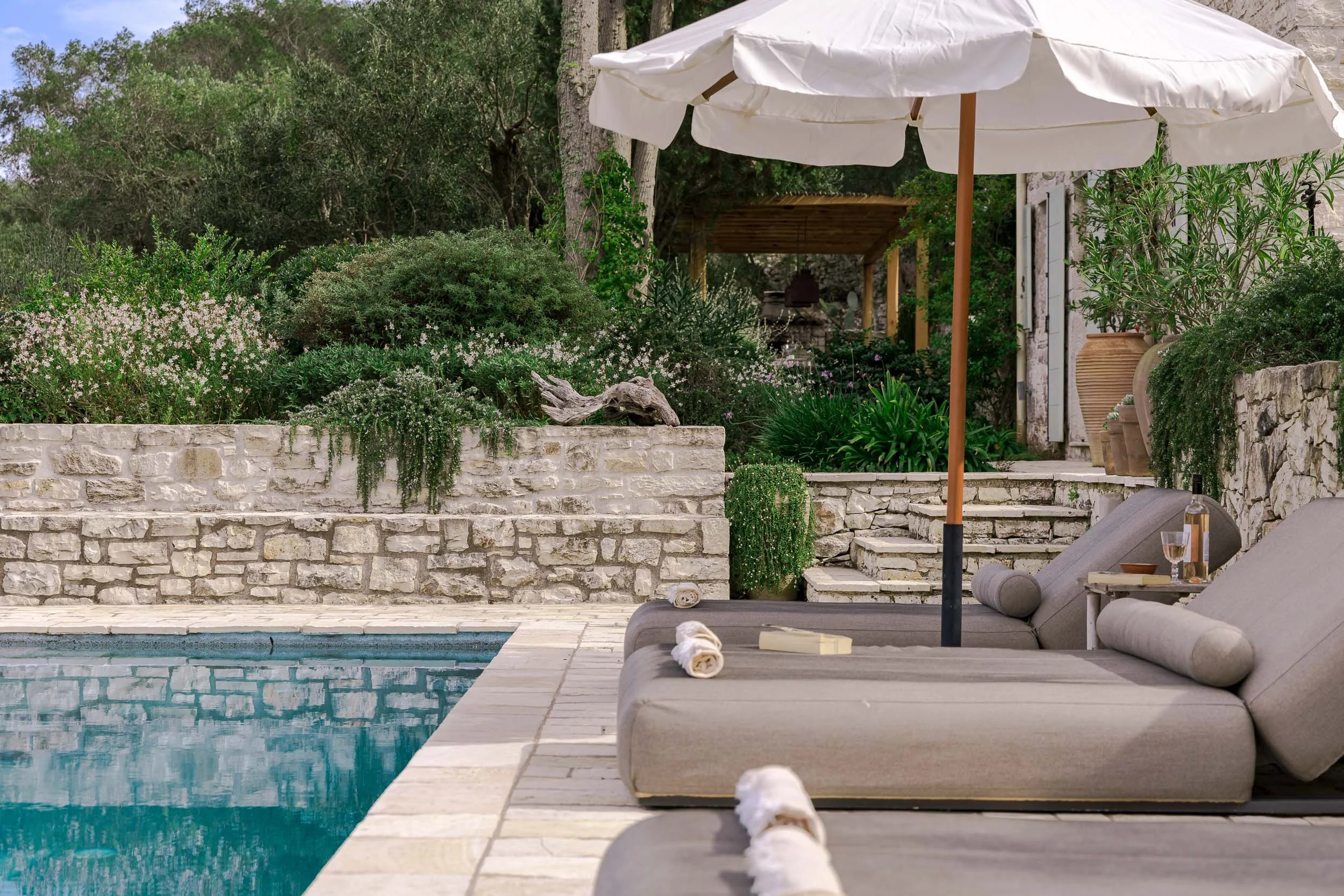 A relaxing outdoor poolside scene with lounge chairs, a white umbrella, and lush greenery in the background.