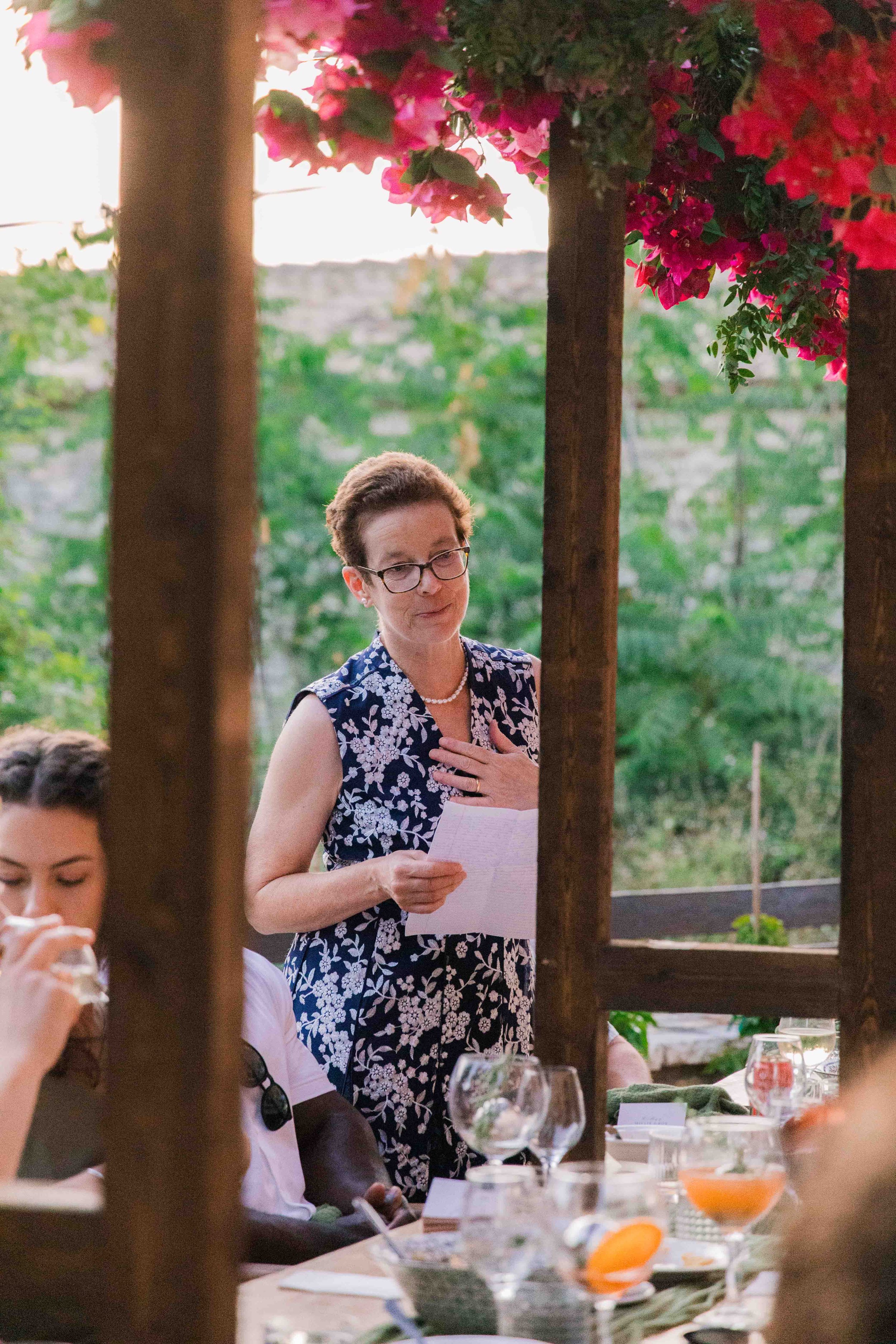 A woman in glasses wearing a blue floral dress is standing and speaking, holding a piece of paper, at a table outdoor gathering surrounded by pink flowering plants and greenery.