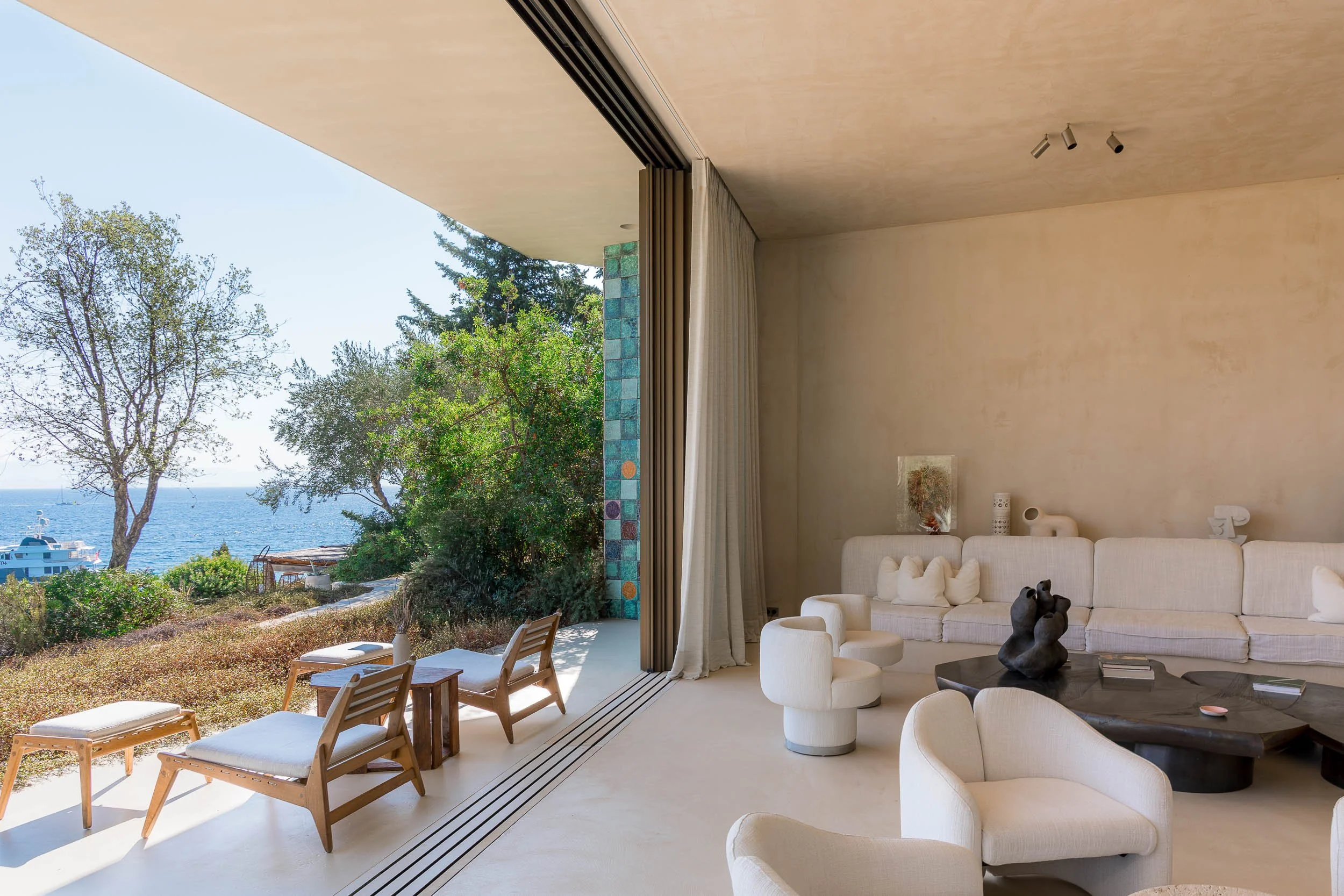 Living room with a large sliding glass door opening to an outdoor patio with chairs, overlooking trees and the ocean.
