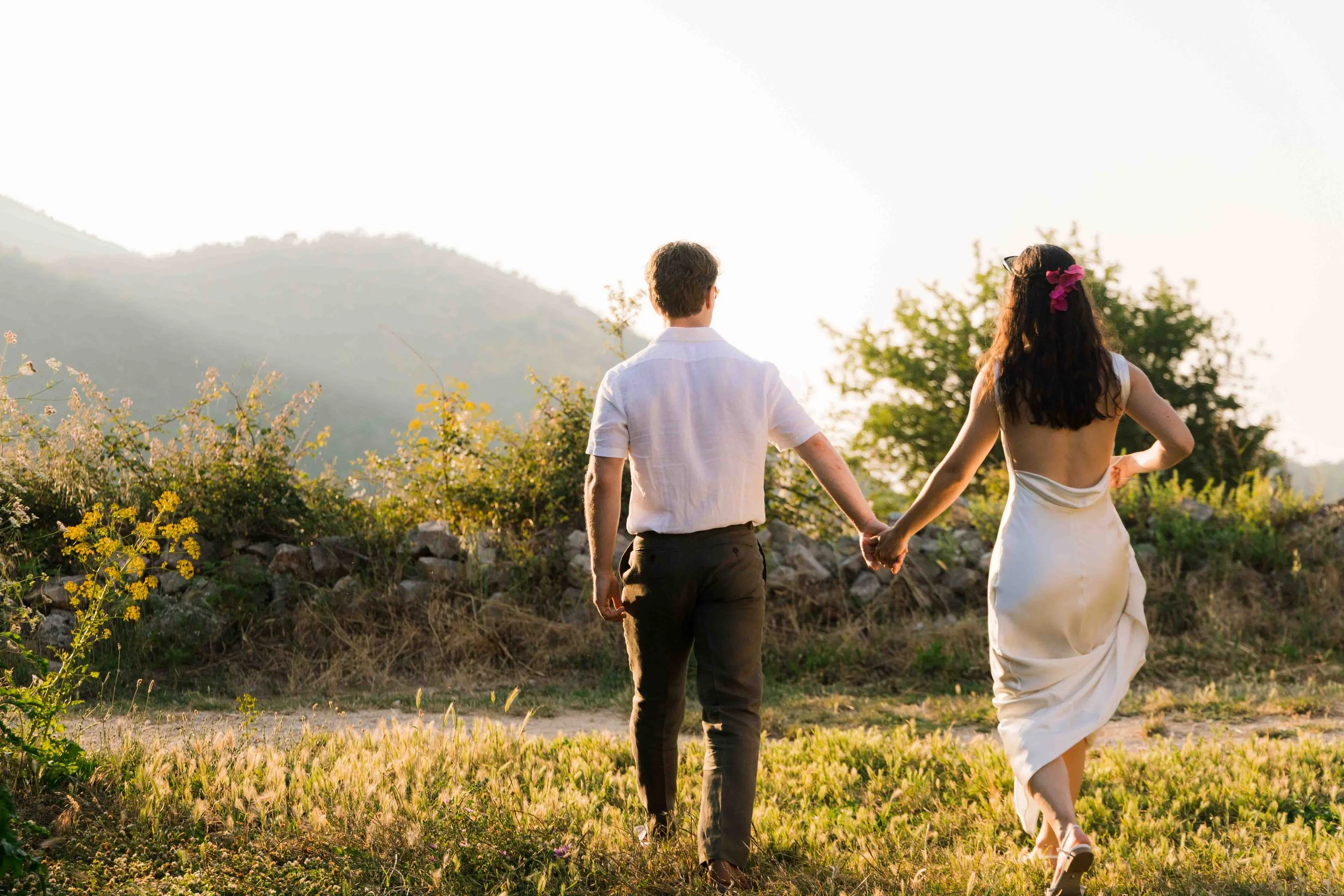 A couple dressed in wedding attire holding hands while walking outdoors during sunset in a rural landscape