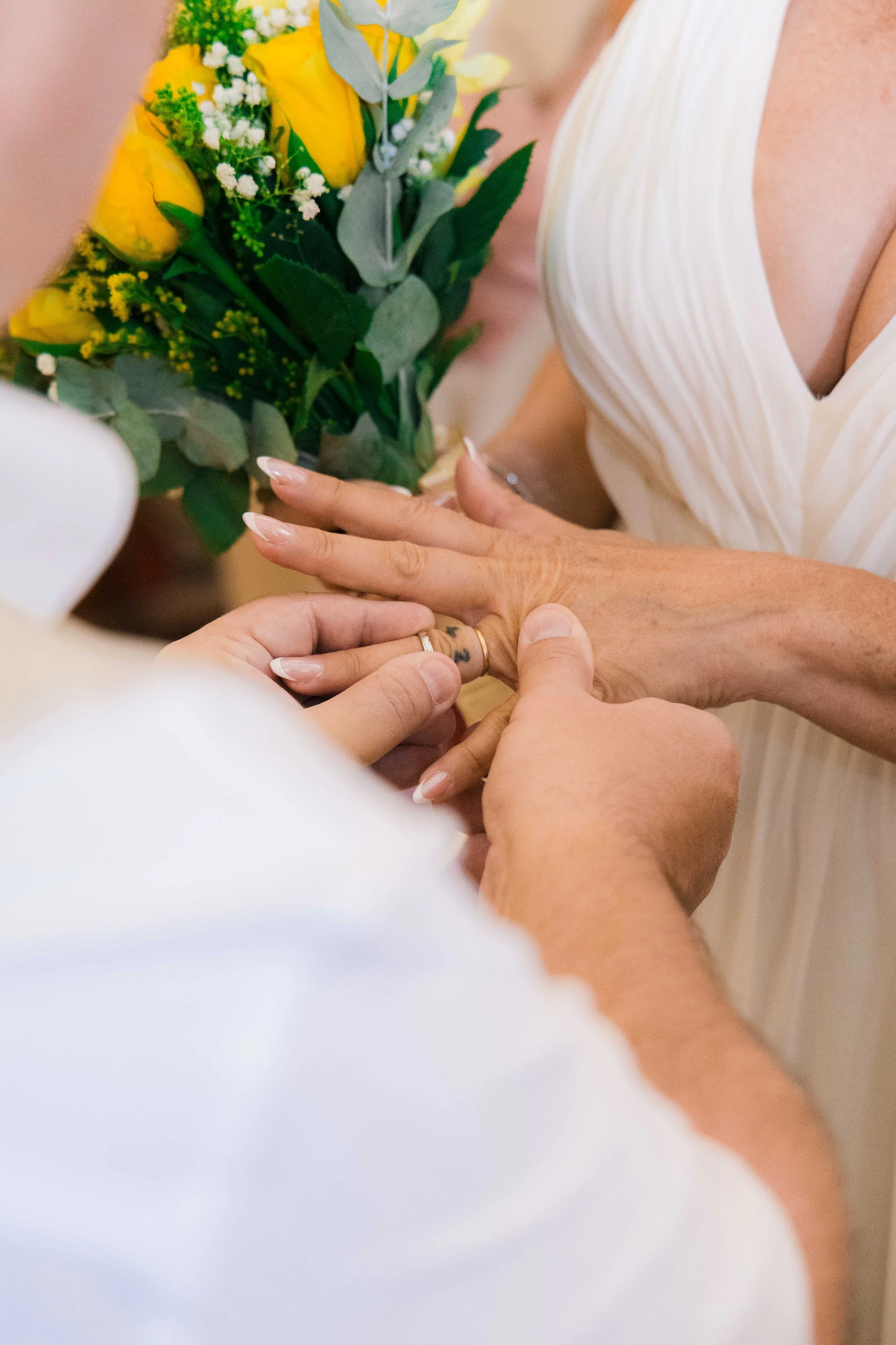 A person is placing a ring on an elderly woman's finger while holding her hand, with a bouquet of yellow flowers in the background.