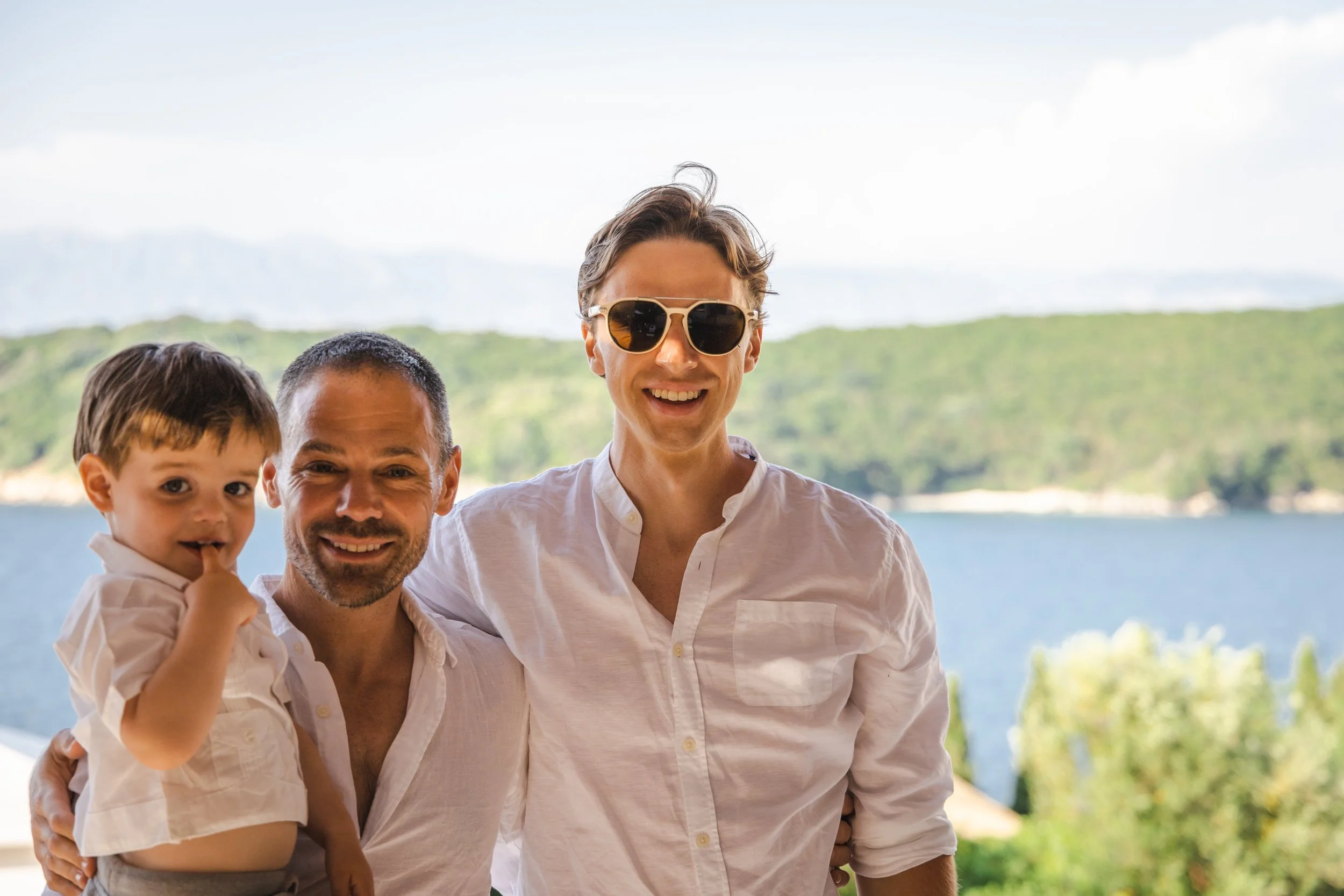Three smiling people — a man, woman, and young boy — standing outdoors near a body of water with a green hillside in the background.