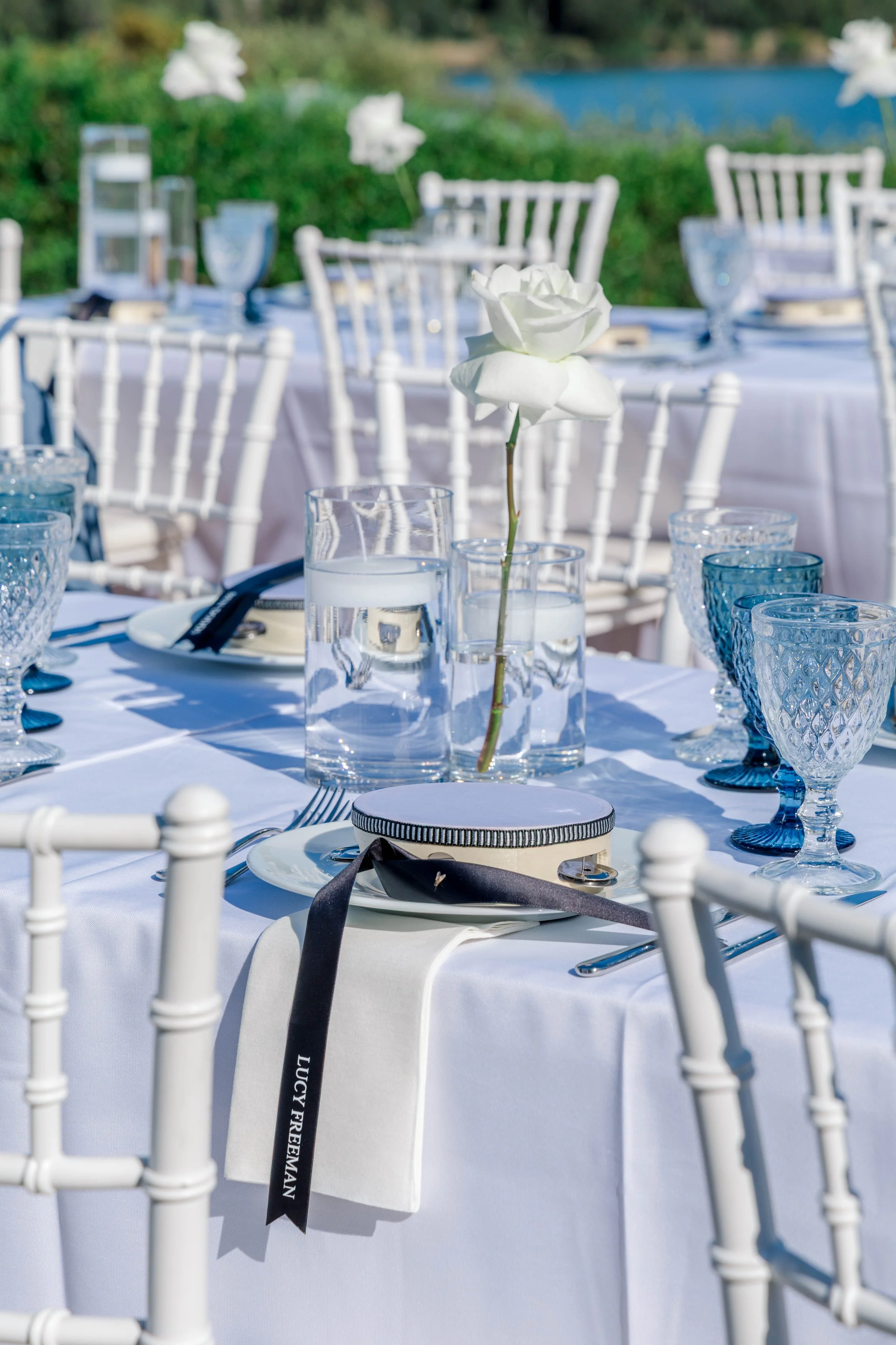 Elegant outdoor table setting with white tablecloth, white chairs, a tall white rose in a glass vase, water glasses, and cutlery, set for a formal event.