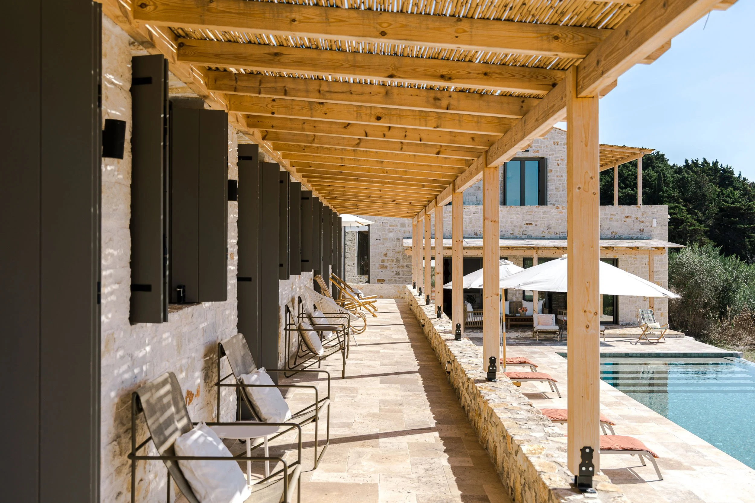 Outdoor patio with lounge chairs and umbrellas next to a swimming pool, stone building, and lush greenery in the background.