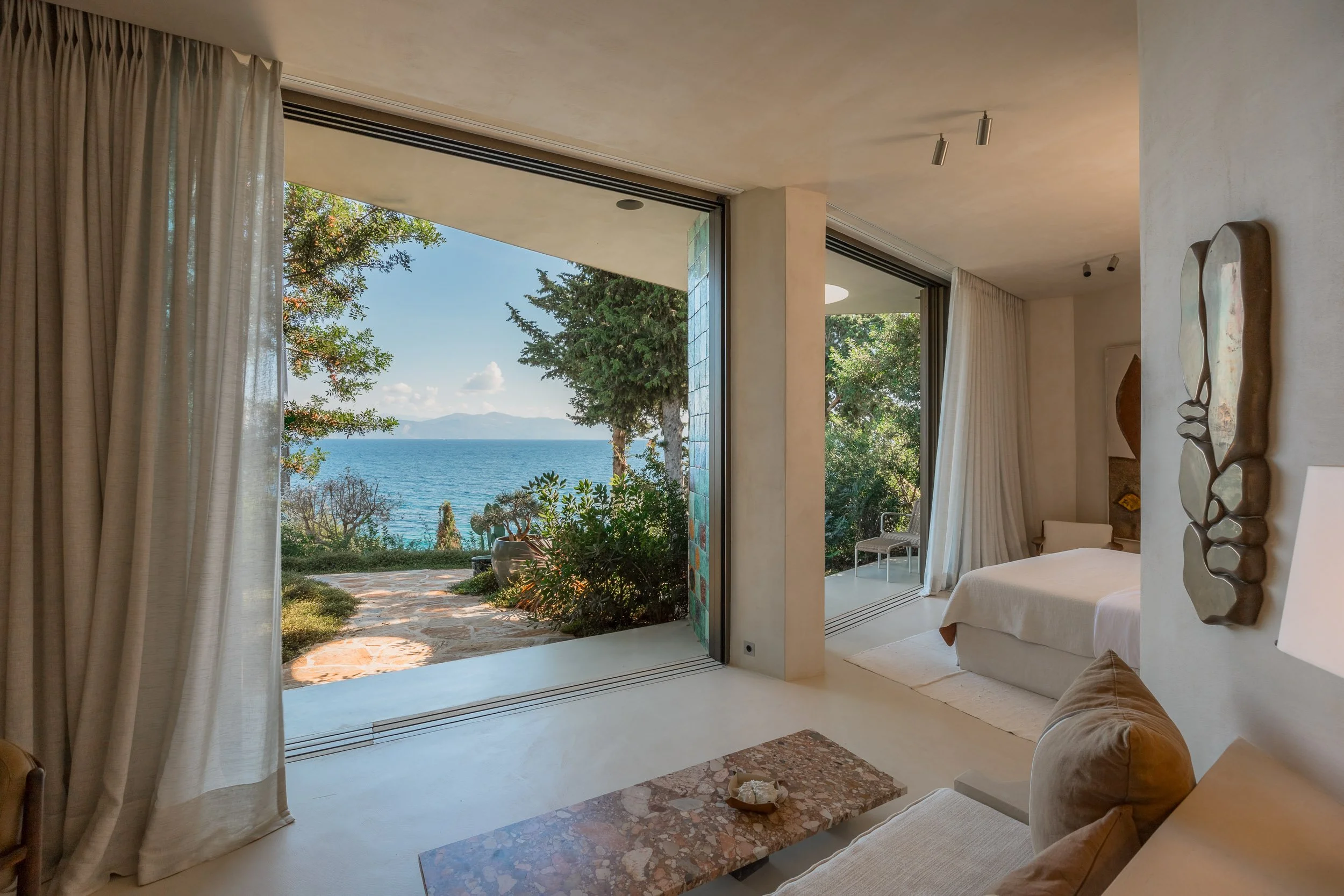 A room with a view of the ocean through large sliding glass doors, leading to a patio with plants and trees.