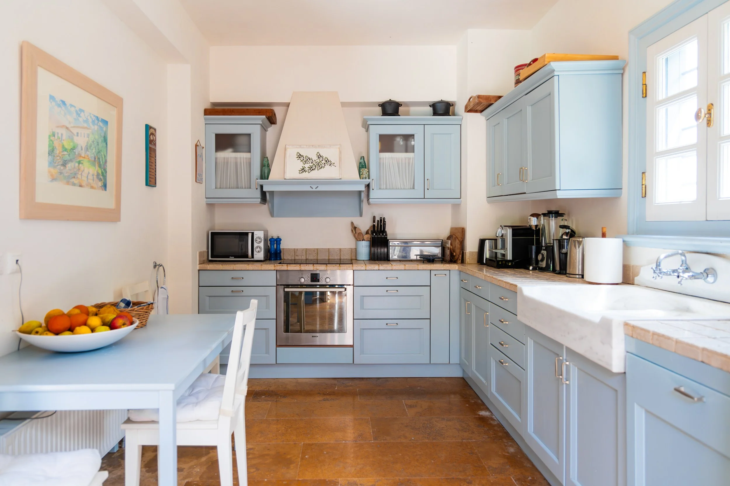 Cozy kitchen with light blue cabinets, beige countertops, a white farmhouse sink, and a window. A small white dining table with a bowl of fruit is in the foreground.