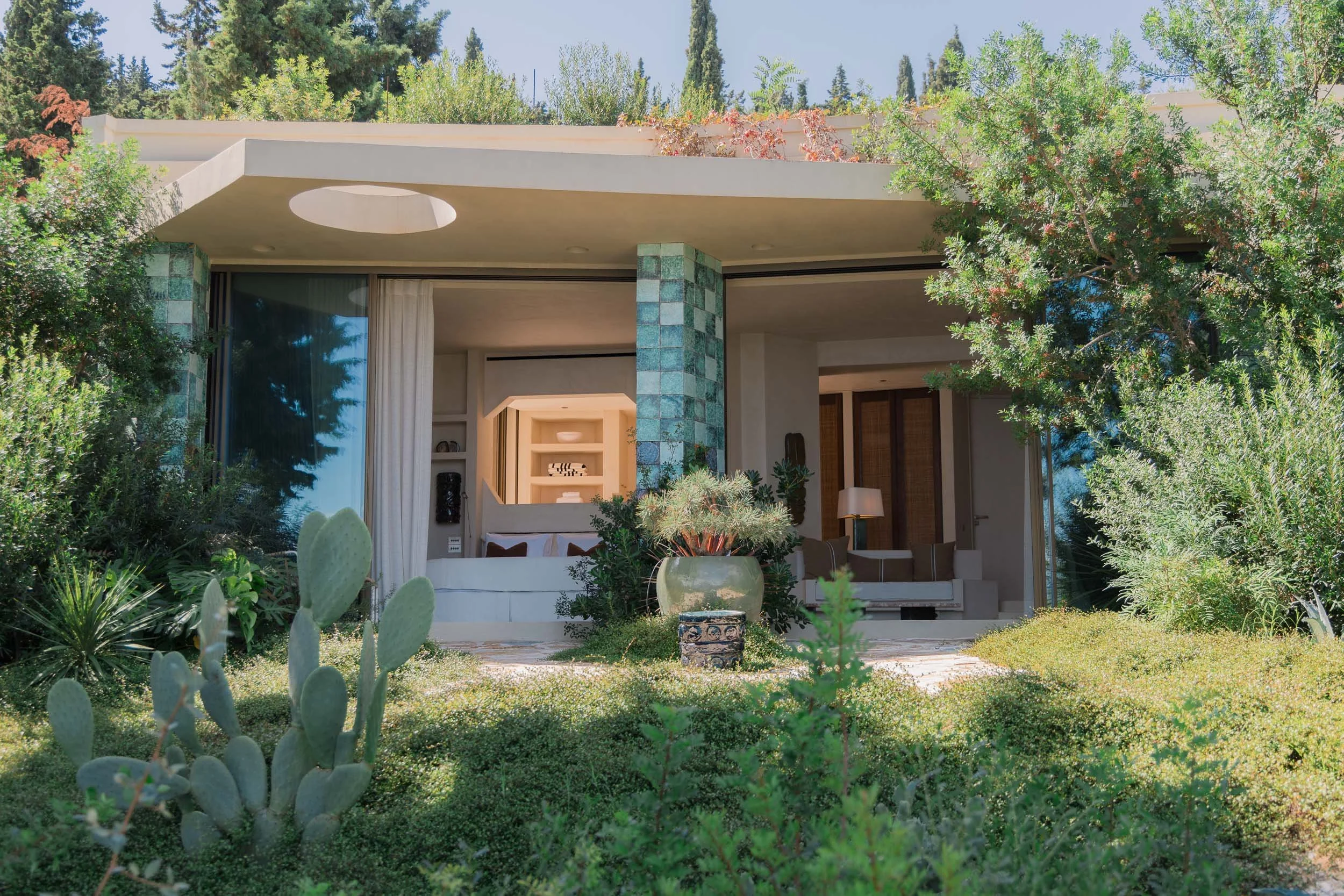 Modern house with an open front porch, surrounded by lush greenery and desert plants, including cacti and bushes, with trees in the background.