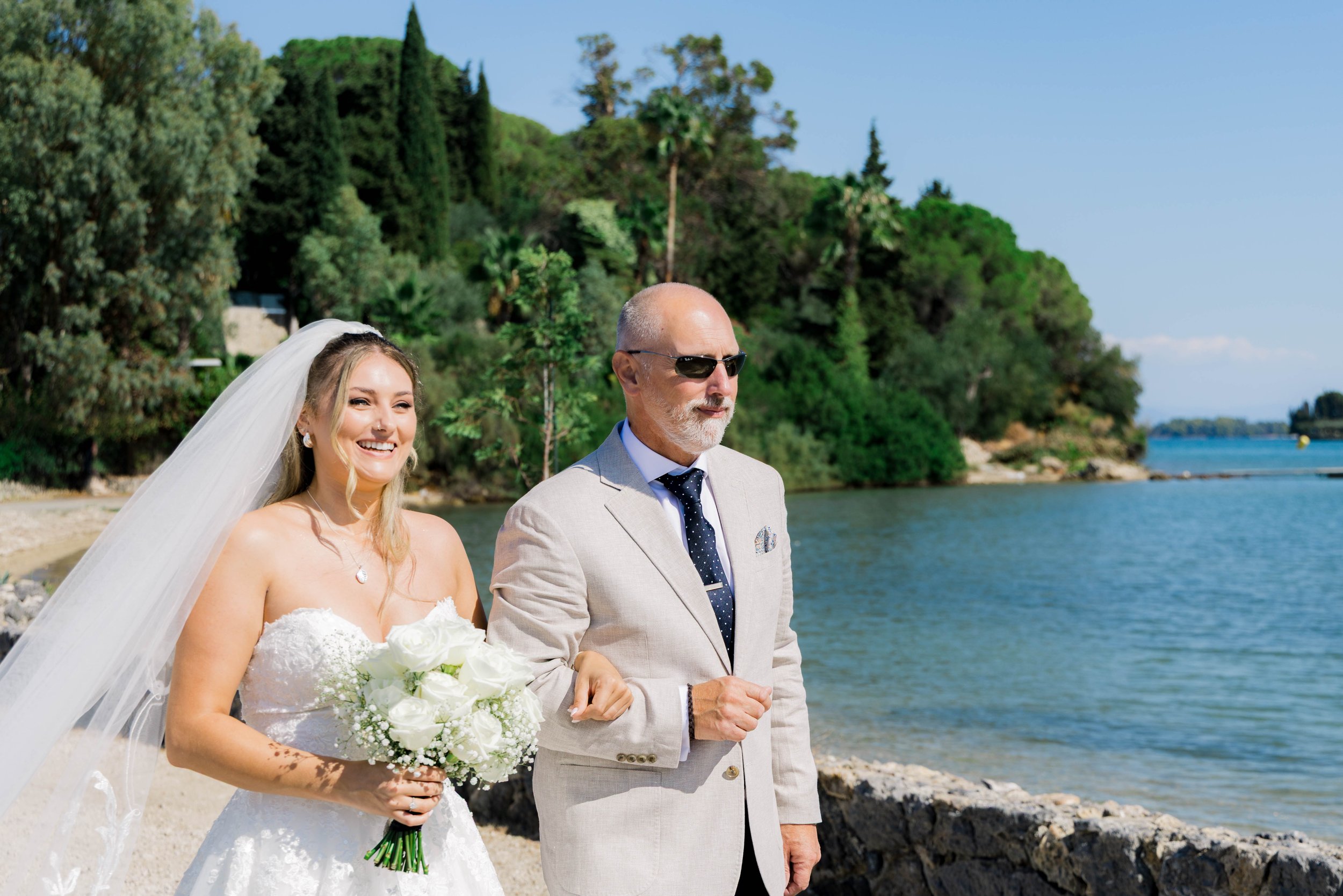A bride in a white wedding dress holding a bouquet of white roses and standing next to an older man in a beige suit and sunglasses, by a lakeside with green trees and blue sky in the background.