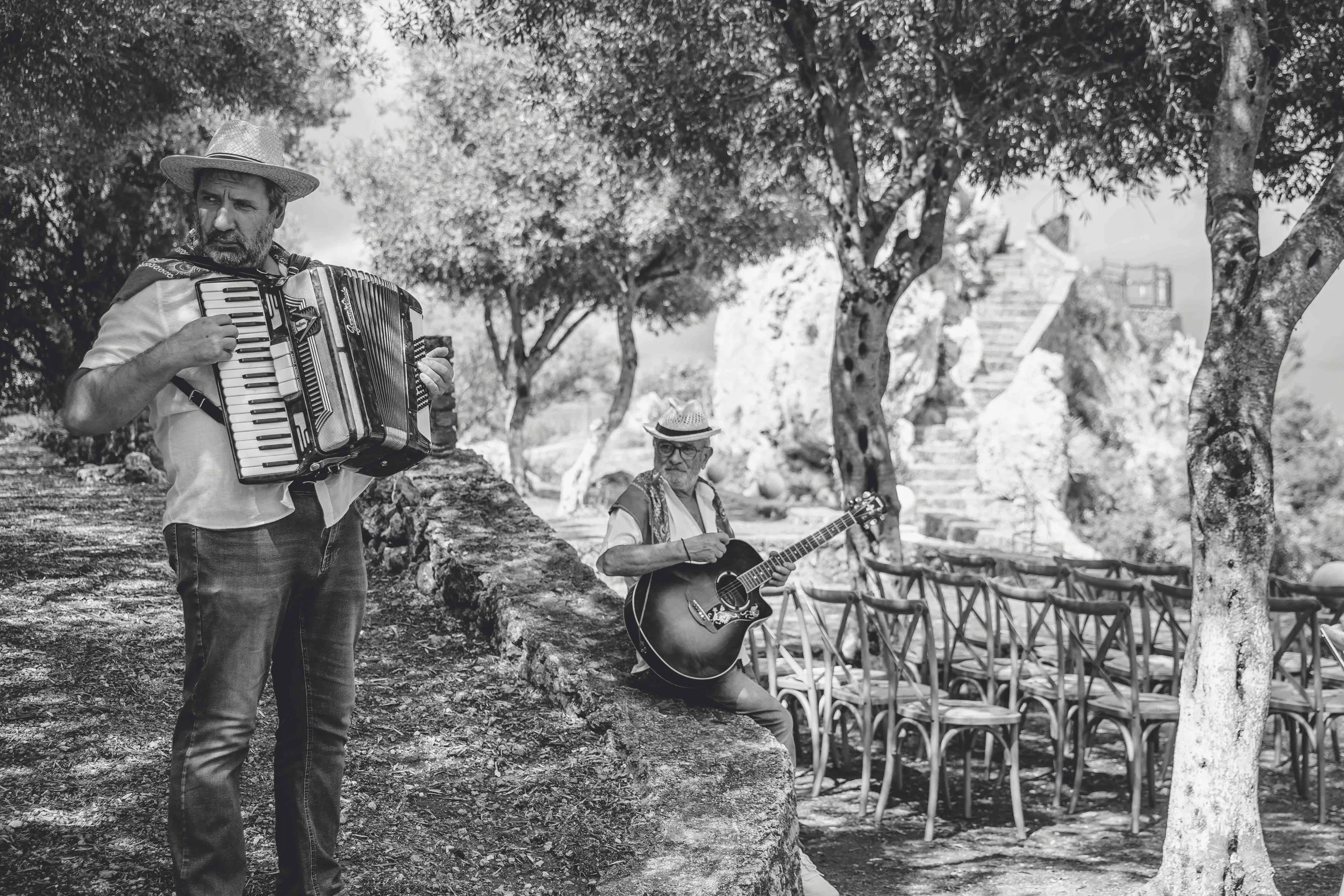Two musicians, one playing an accordion and the other playing an acoustic guitar, perform outdoors among trees and chairs.