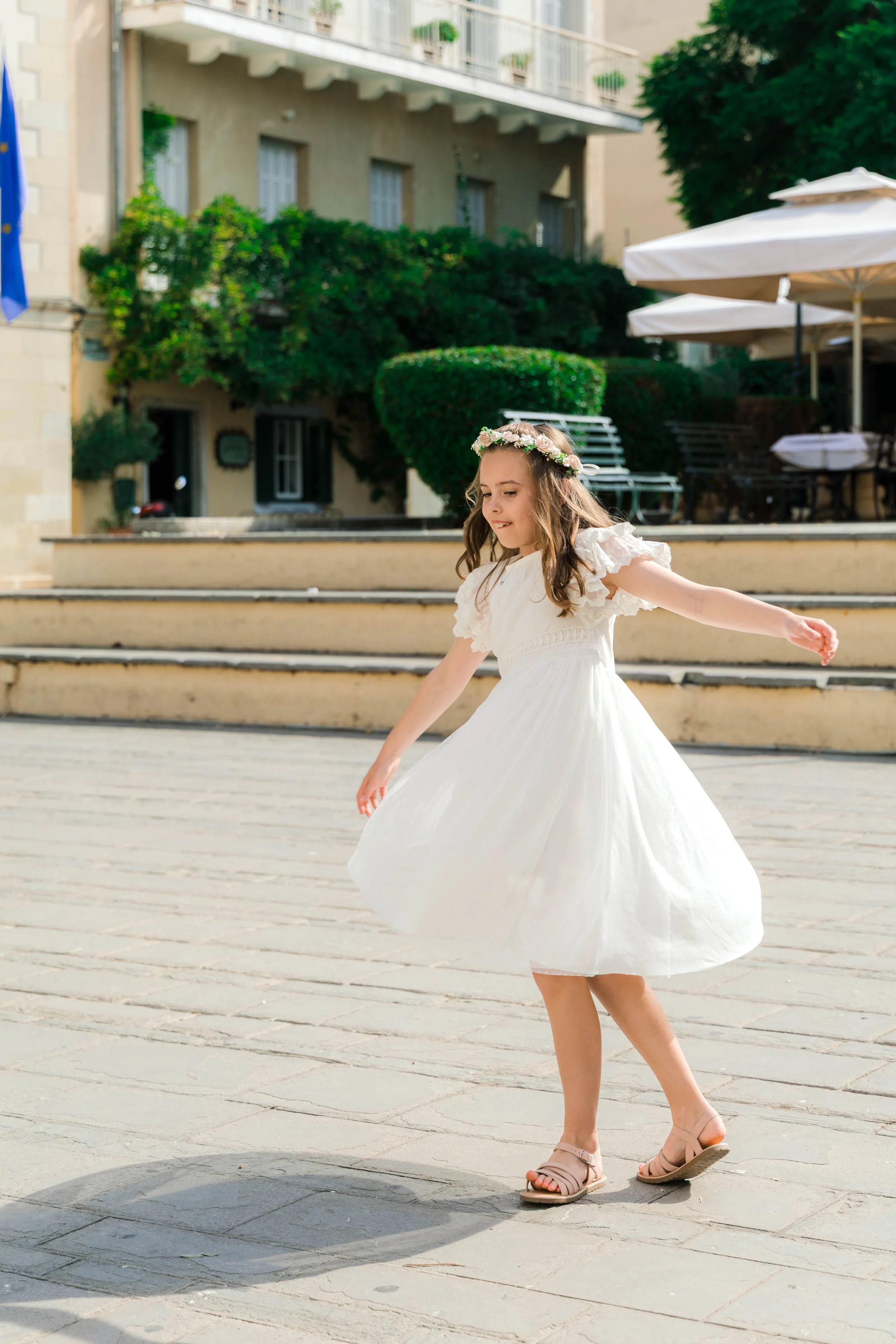 A young girl in a white dress spinning around outside on a paved area, wearing a floral crown and beige sandals.