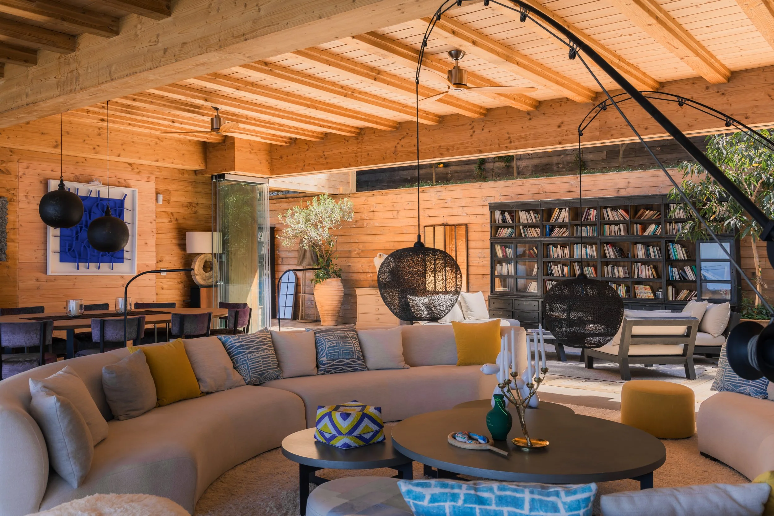 A spacious living room with wooden walls and ceiling, featuring a large beige sectional sofa with colorful pillows, a round coffee table with decorative items, black hanging light fixtures, a bookshelf filled with books, a dining table with chairs, a