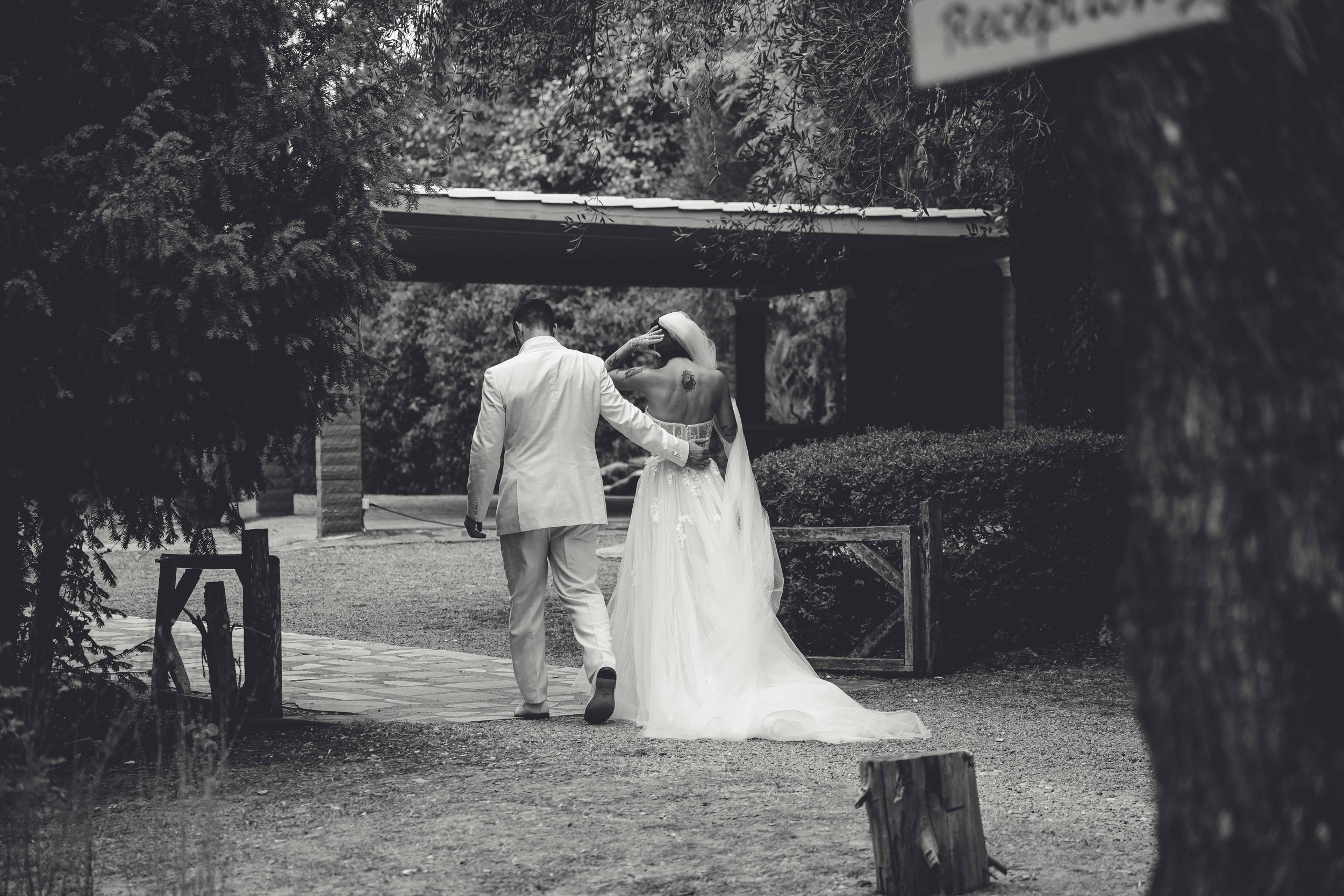 A black and white photo of a couple in wedding attire walking outdoors, surrounded by trees and bushes.