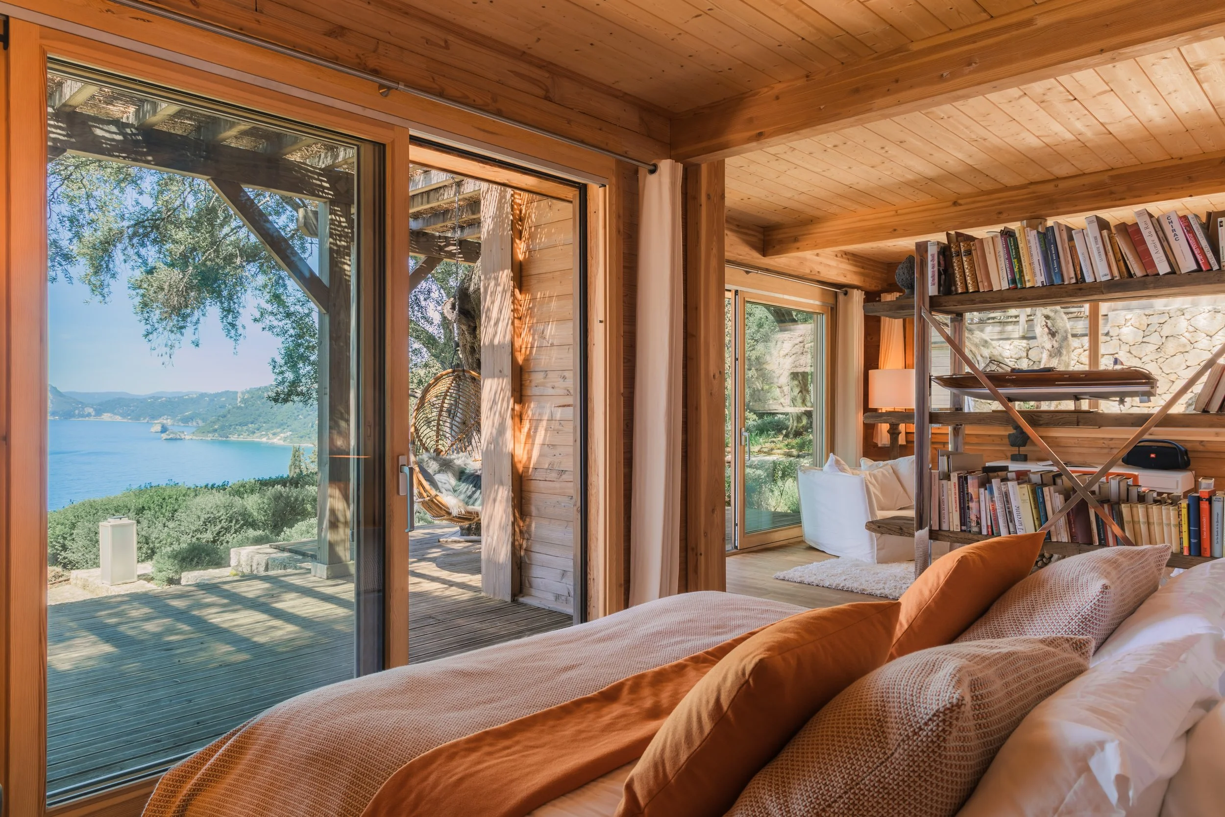A cozy bedroom with wooden walls and ceiling, large glass sliding doors leading to a deck with a view of a lake and mountains, a bookshelf, and a sitting area with white furniture.