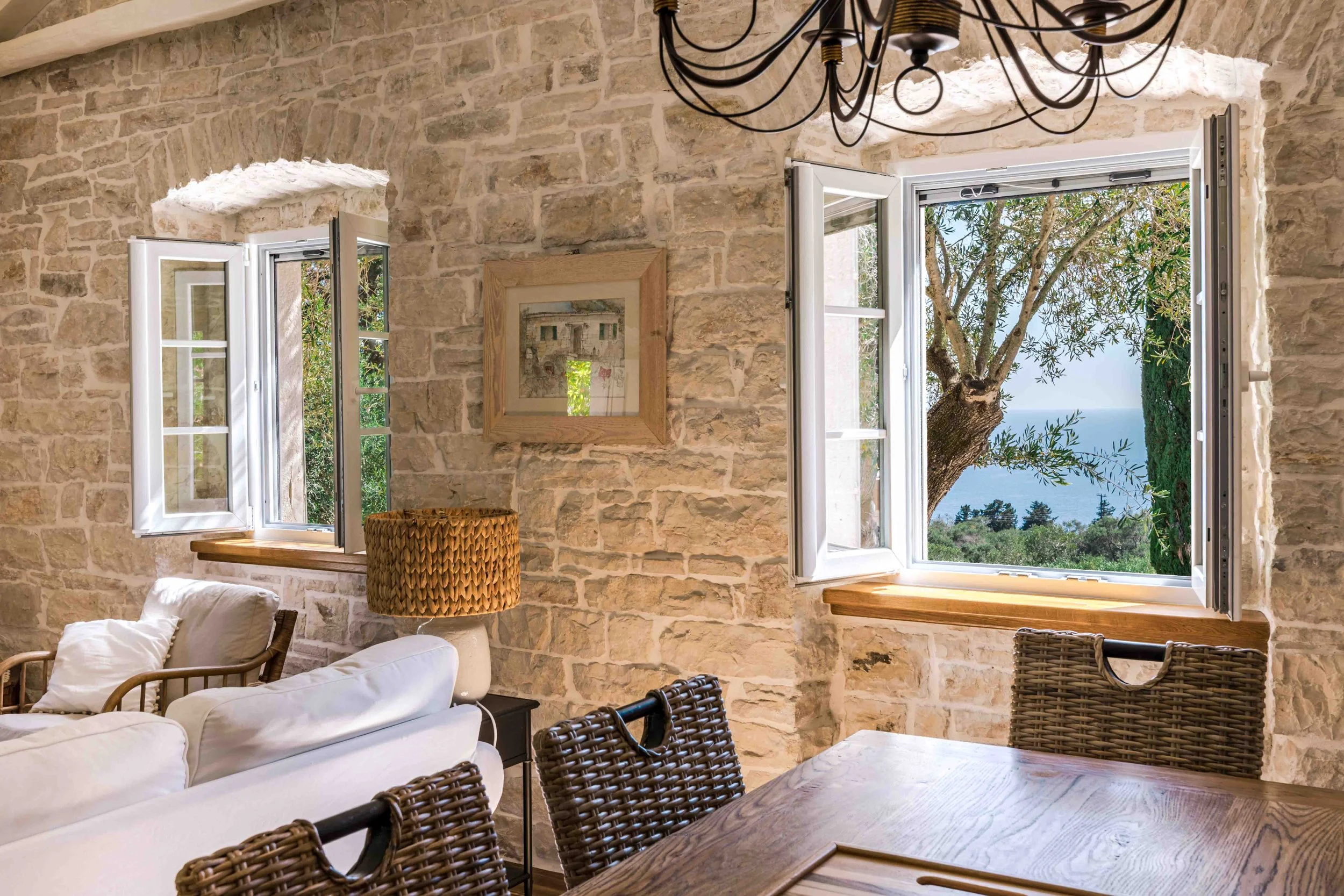 Interior of a room with stone walls, two open windows showing a tree and view of the ocean, a wooden dining table with woven chairs, a white sofa with pillows, a lamp with a woven shade, and framed artwork on the wall.