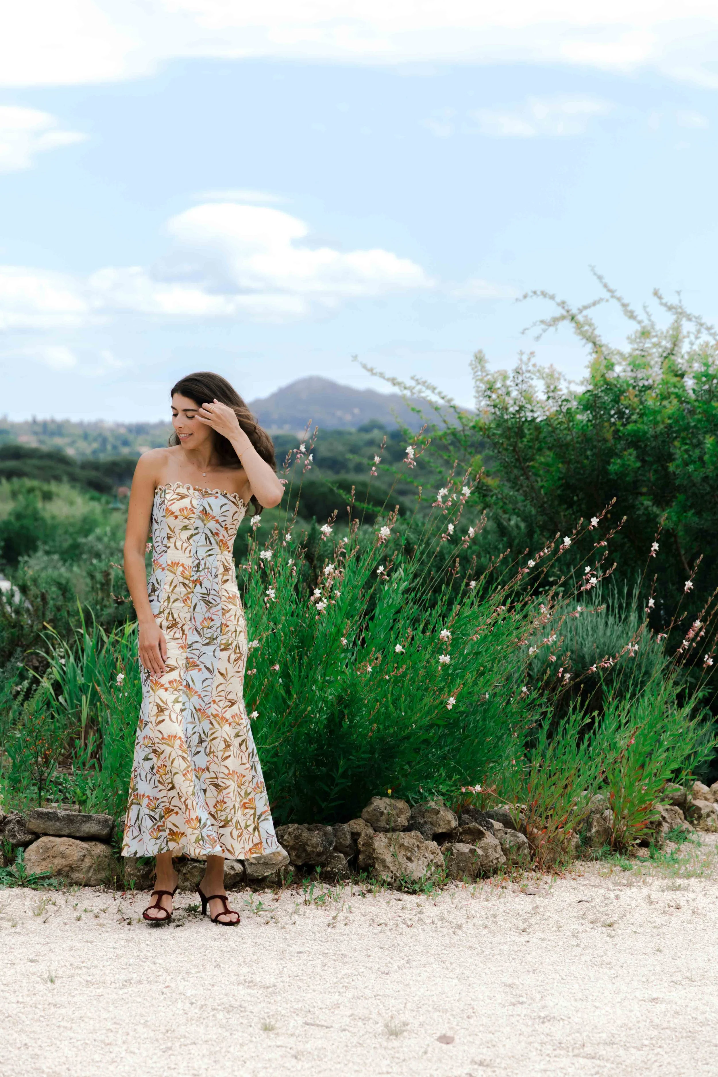 Woman in a strapless floral dress standing outdoors in front of greenery and rocks, smiling and touching her hair.