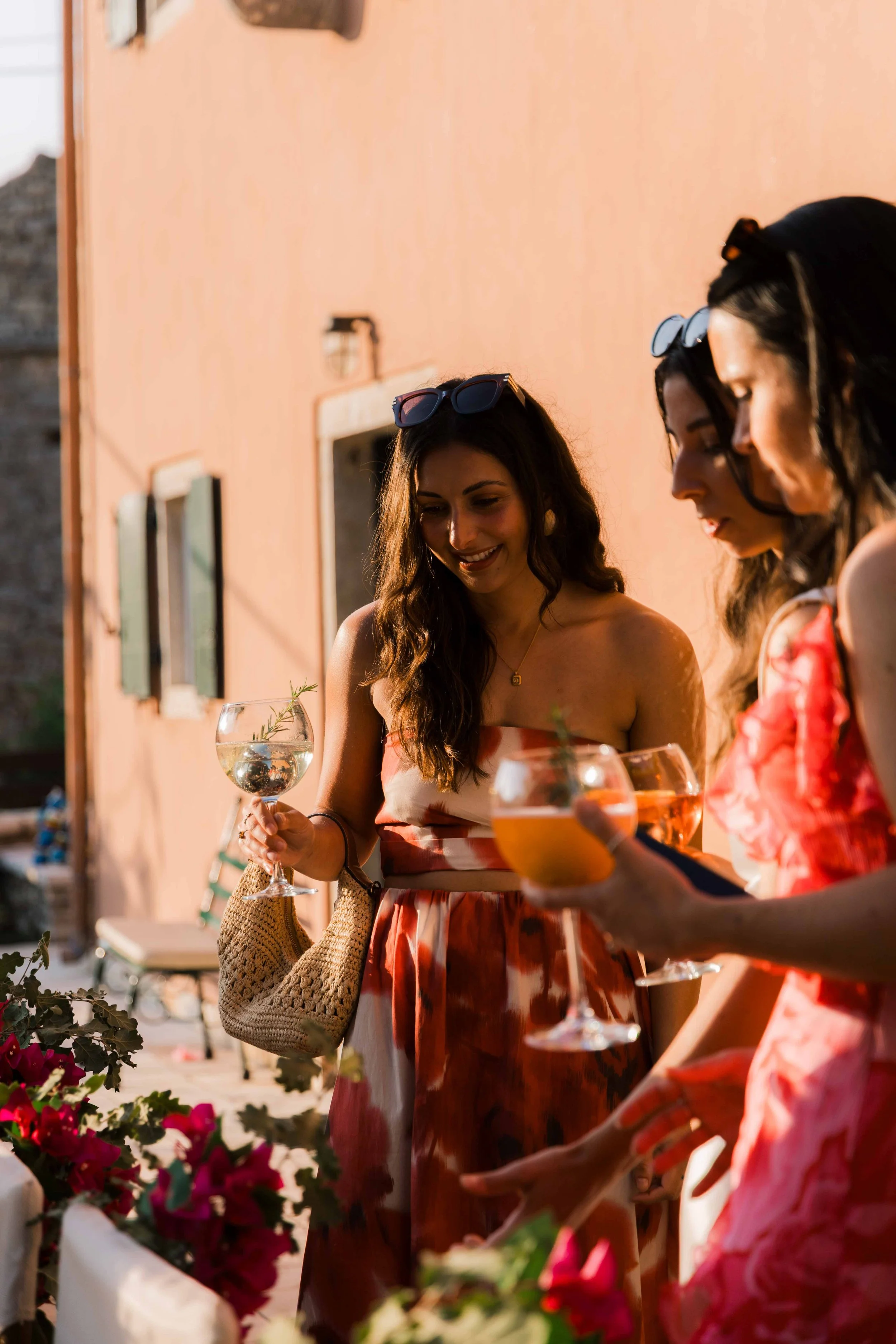 Three women enjoying drinks outdoors, standing near pink flowers and a coral-colored building, wearing summer dresses, with sunlight enhancing the scene.