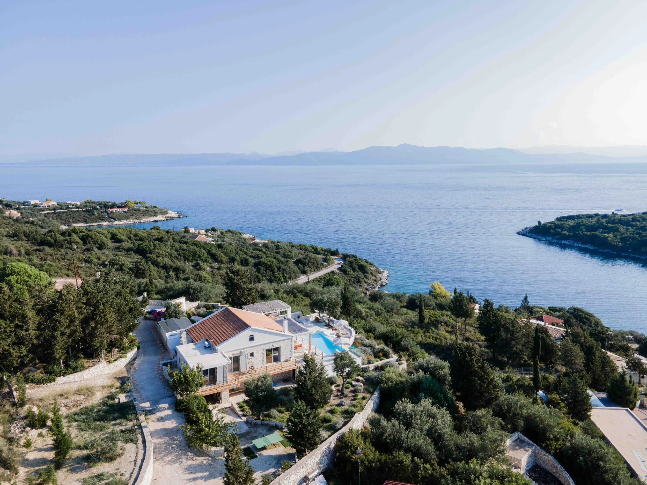 Aerial view of a coastal landscape showing a modern house with a swimming pool, surrounded by lush greenery, overlooking a large body of water with islands or land masses in the distance.