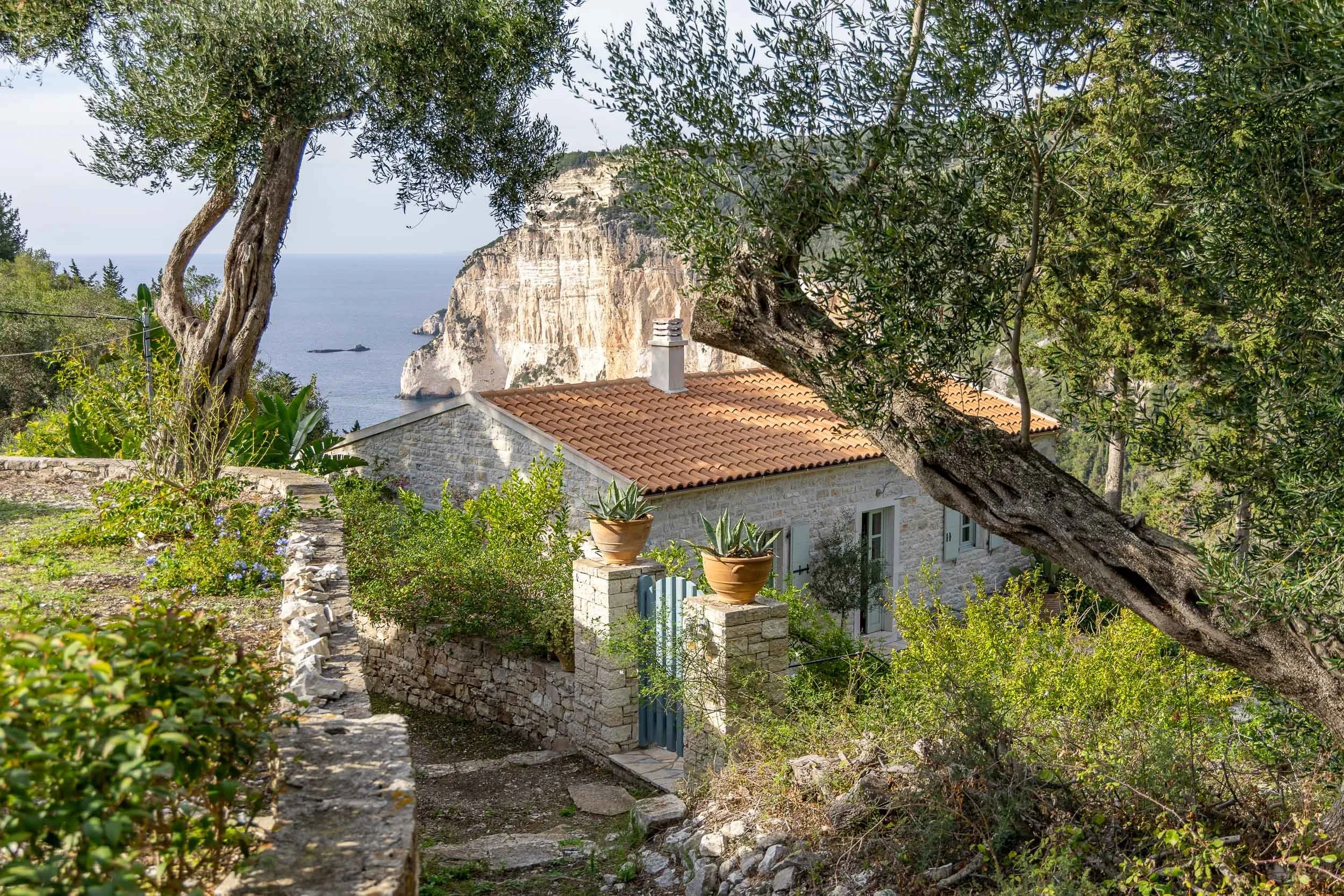 A stone house with a red tiled roof, surrounded by trees and greenery, with a view of cliffs and the sea in the background.