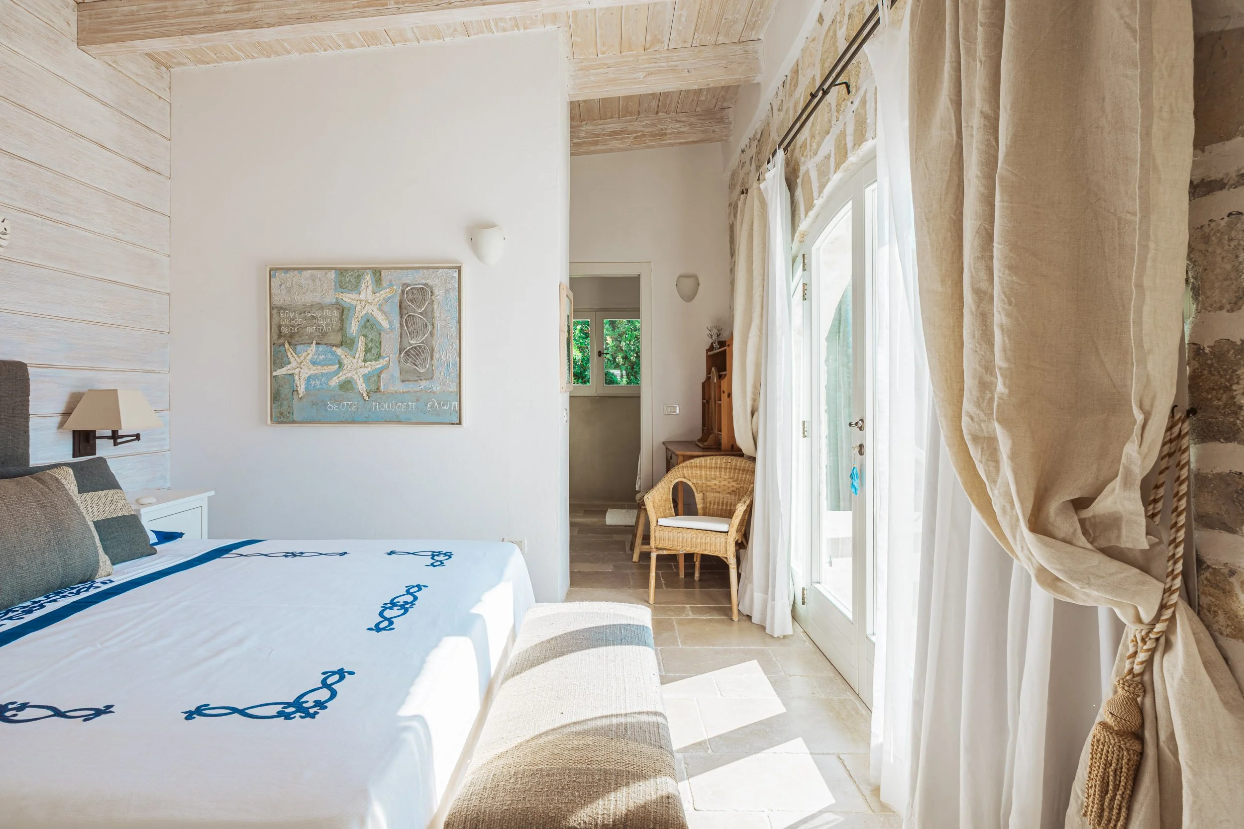 Interior view of a bright bedroom with a white bed, beige and gray pillows, wooden panel wall, a piece of artwork on the wall, hanging white curtains on a glass door, a wicker chair, and a small wooden writing desk.