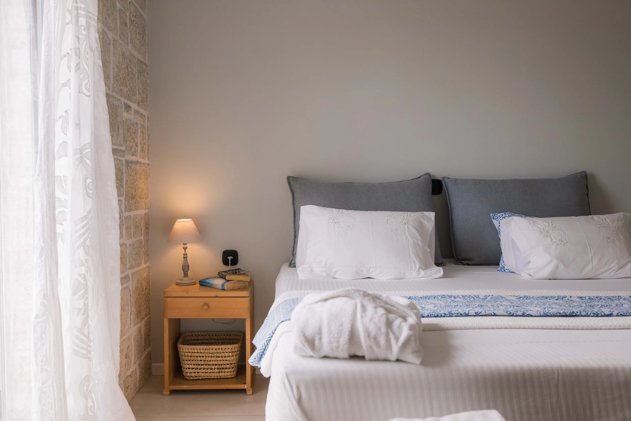 A bedroom with a bed, grey headboard, white linens, and pillows. A small wooden nightstand with a lamp, books, and a basket. A stone wall on the left and white curtains on the window.