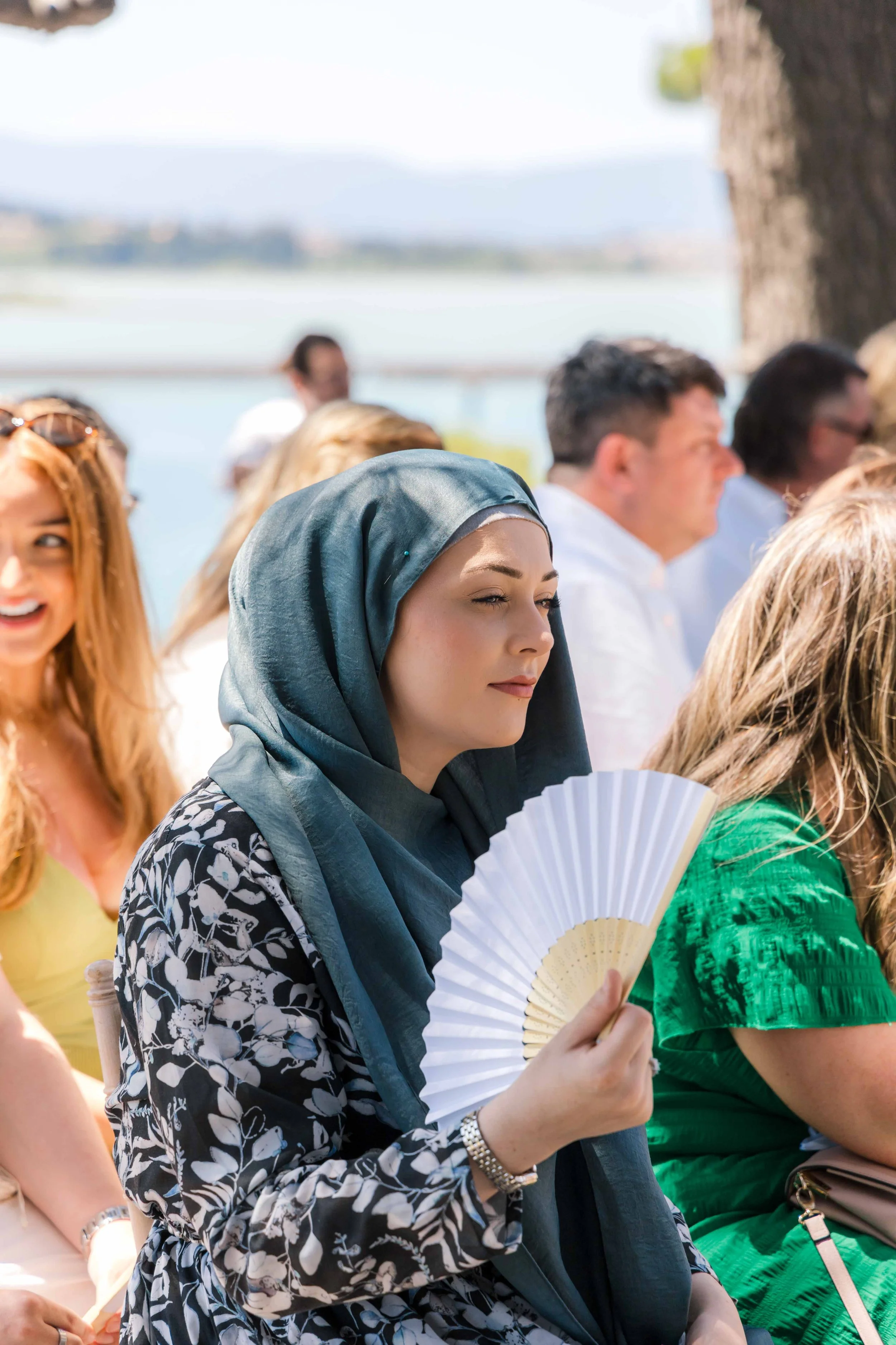A woman wearing a dark headscarf and patterned dress holding a white fan at an outdoor event near a body of water, with other people seated nearby.