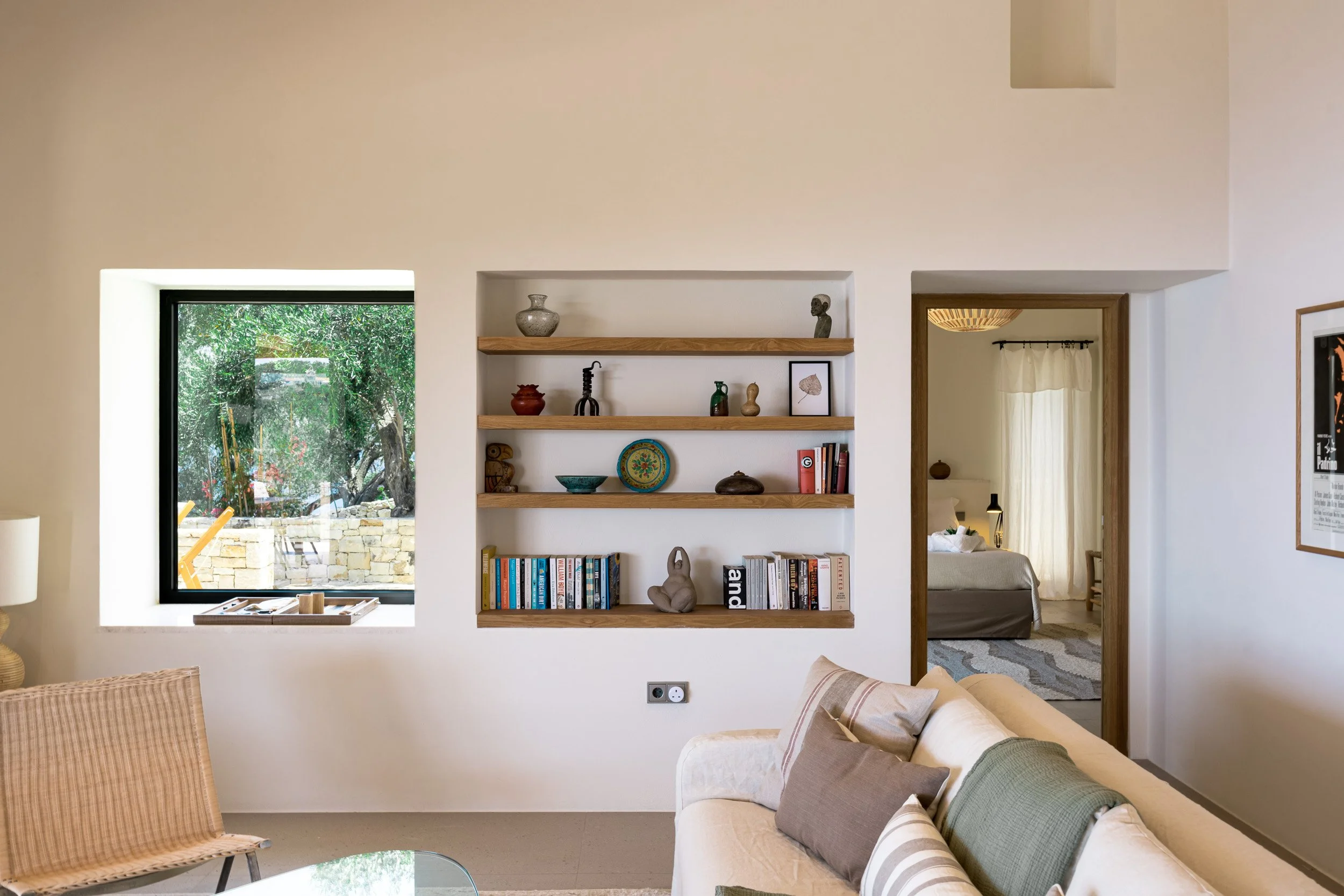 Living room with a window on the left, built-in shelves with decorative items, and a doorway to a bedroom in the background.