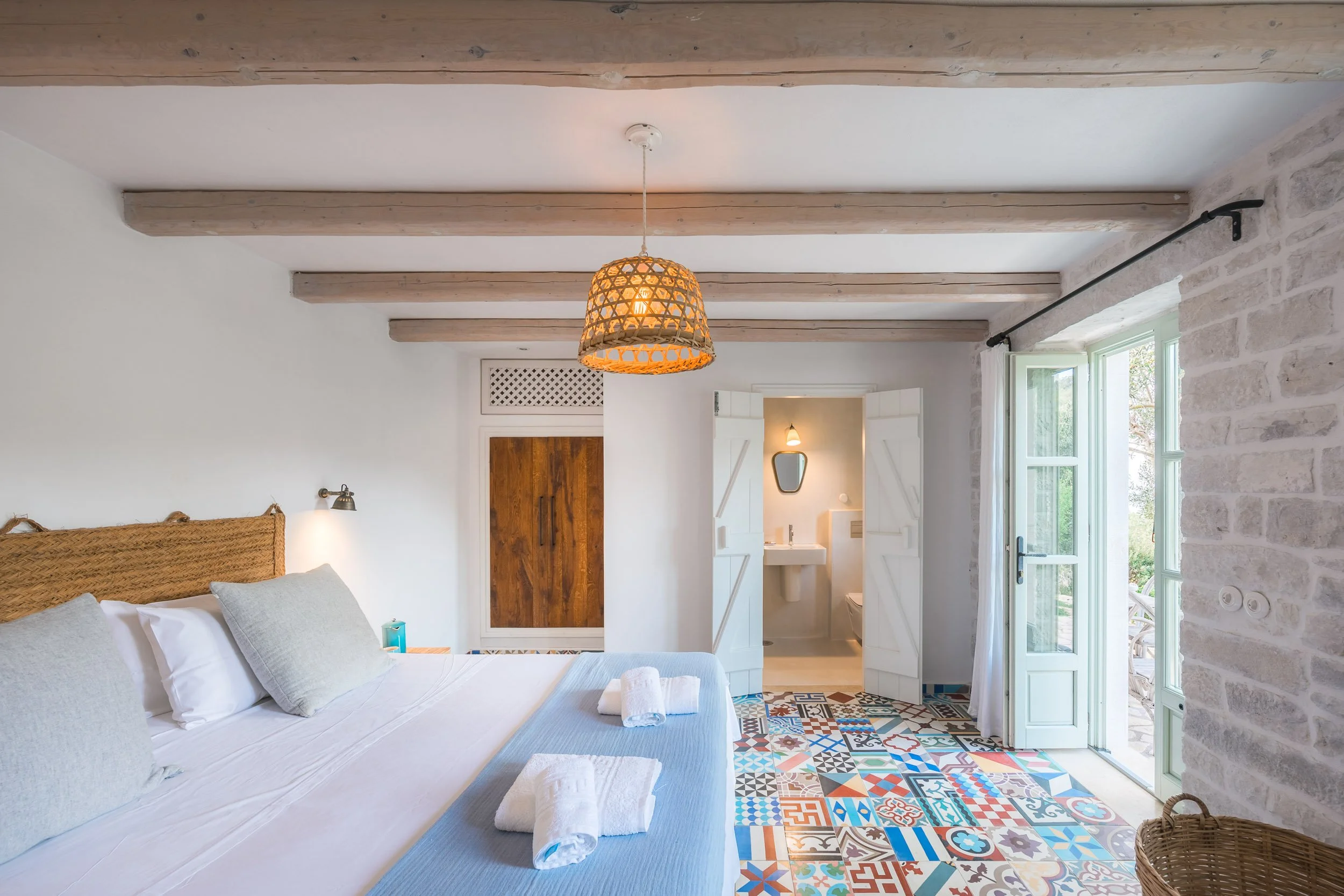 Bright bedroom with a large bed, light-colored pillows, and folded towels. The floor has colorful patterned tiles. There is a door leading to a balcony or patio outside, a small bathroom with a sink and mirror visible through open white shutters, and