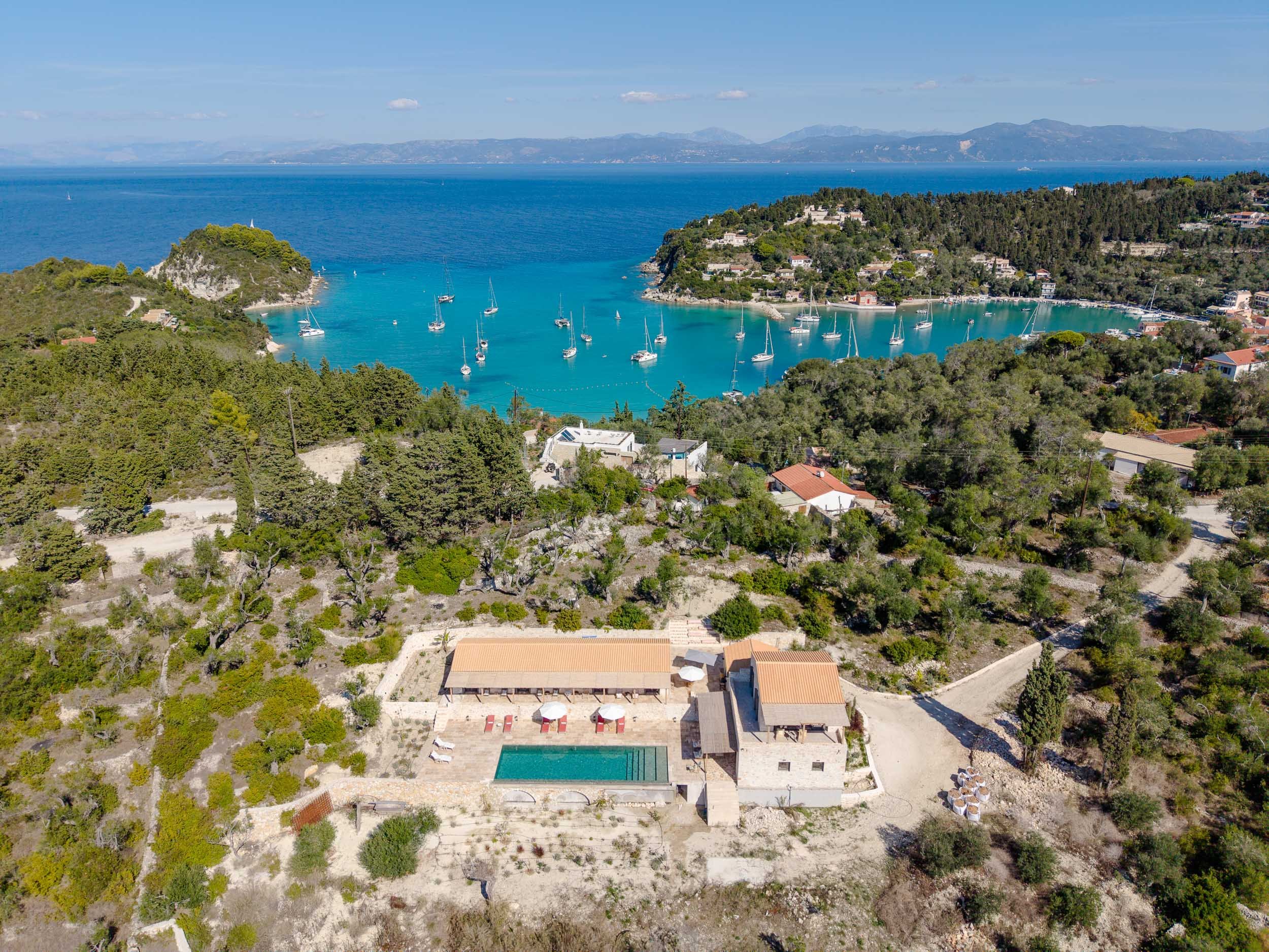Aerial view of a coastal area with a bay containing sailboats, surrounded by lush green hills and houses. In the foreground, there is a property with a swimming pool, patio, and buildings.