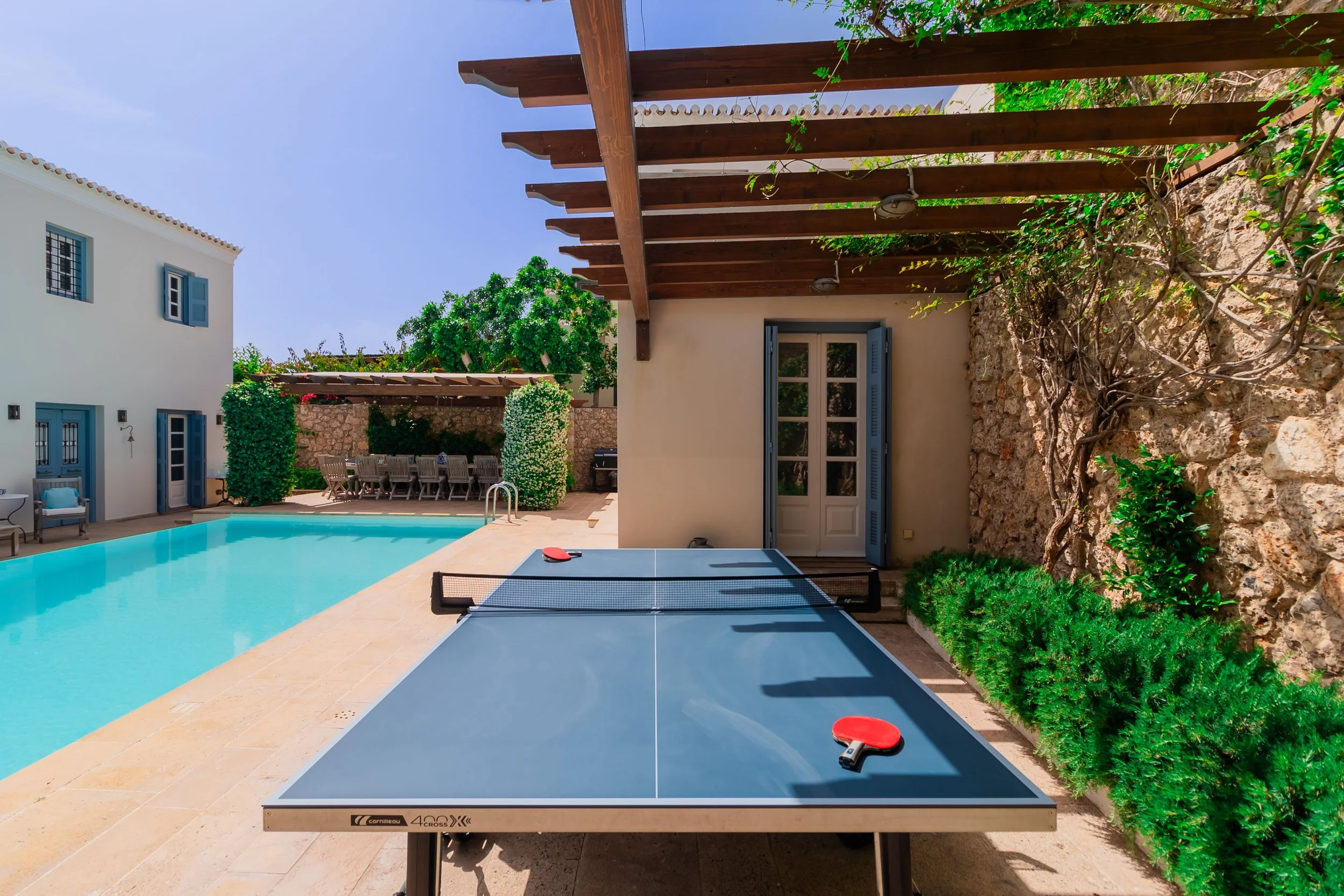Outdoor patio with swimming pool, table tennis table with paddles, grassy plants, stone walls, and a covered seating area under a wooden pergola.
