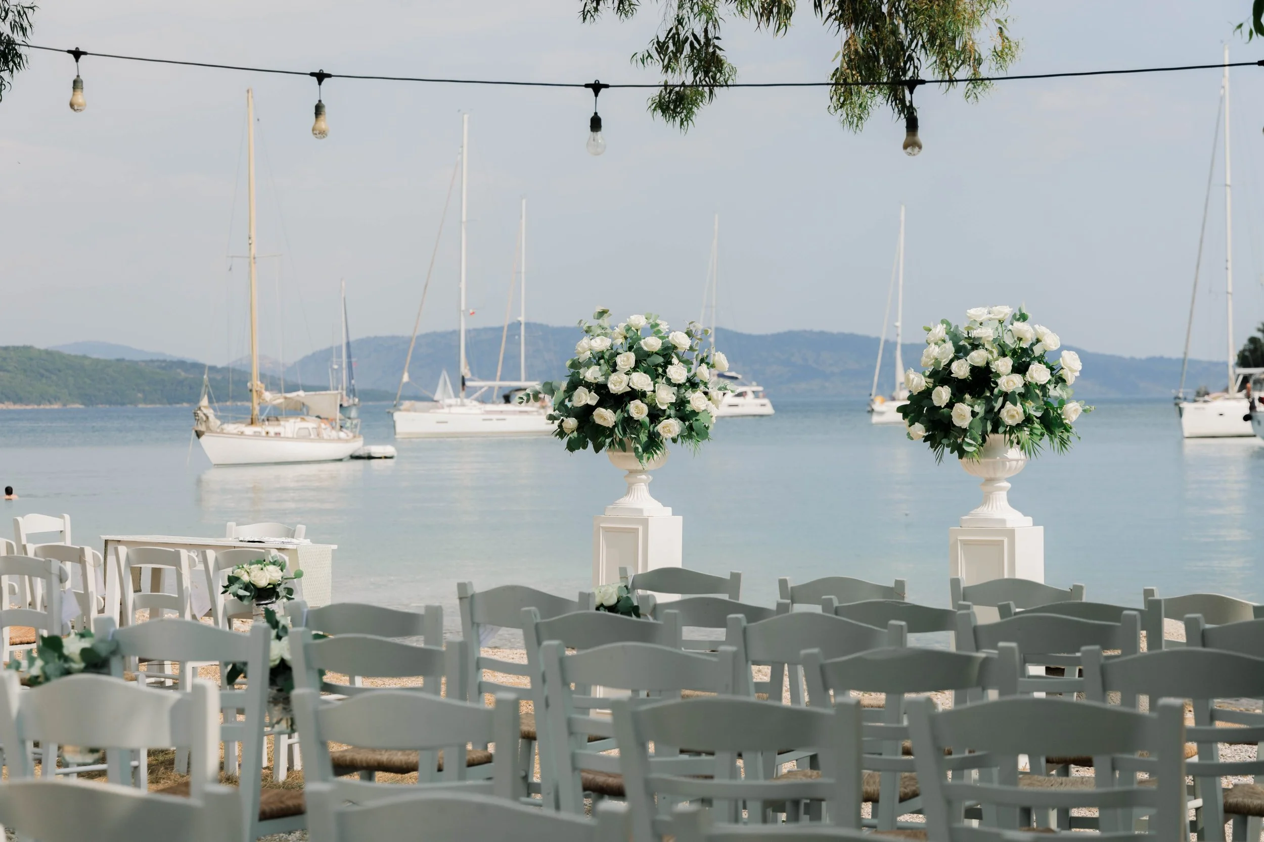 Wedding setup overlooking a lake with sailboats, white chairs, and large floral arrangements with white roses.