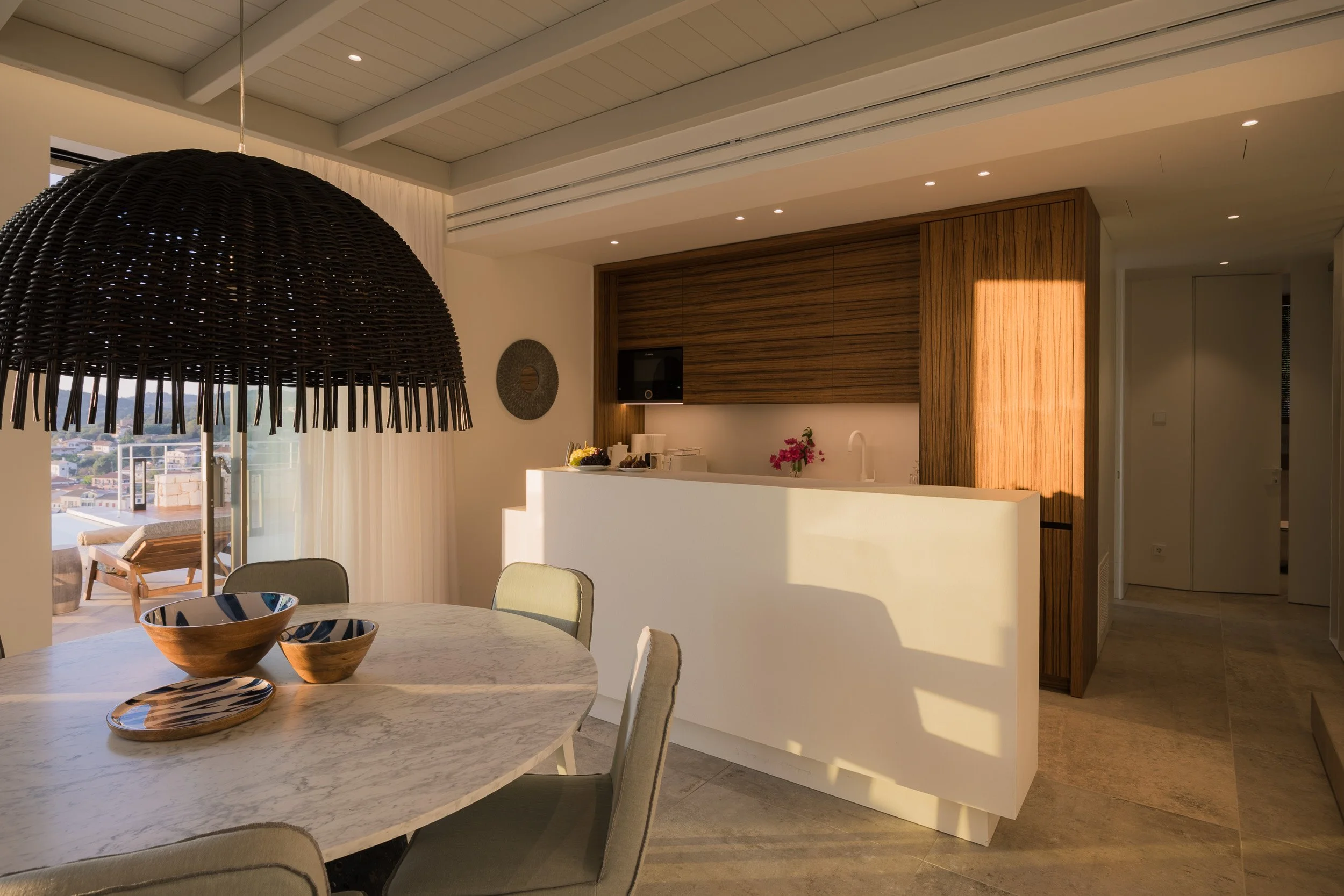 Modern kitchen and dining area with a round marble table, beige chairs, black wicker pendant light, wooden cabinets, and a balcony with outdoor seating, bathed in warm natural sunlight.