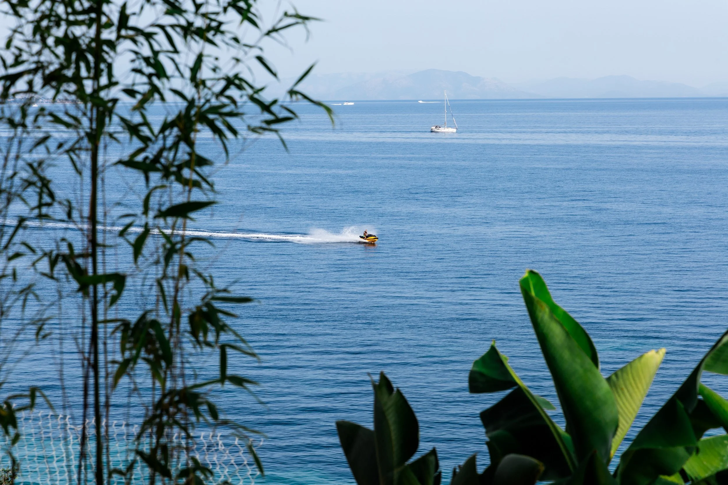 A view of a calm blue lake with a sailboat in the distance and a speedboat creating a wake, framed by green foliage in the foreground and mountains in the background.