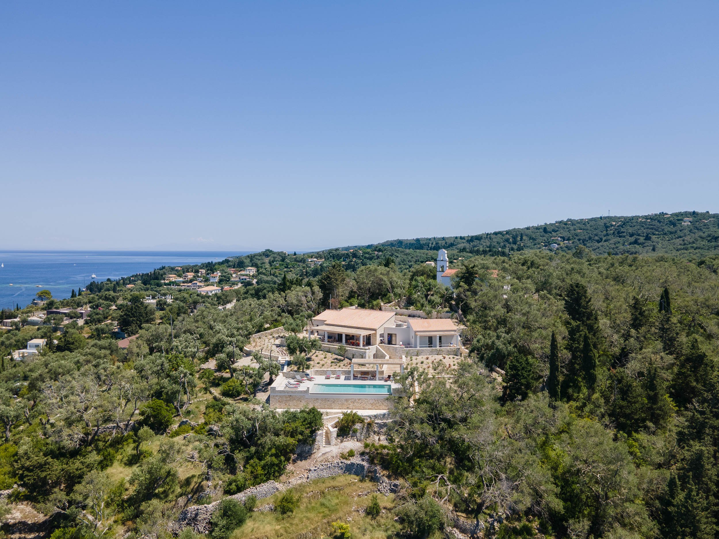 Aerial view of a modern house with a swimming pool, surrounded by trees, overlooking a hilly landscape with other houses, near the ocean under a clear blue sky.