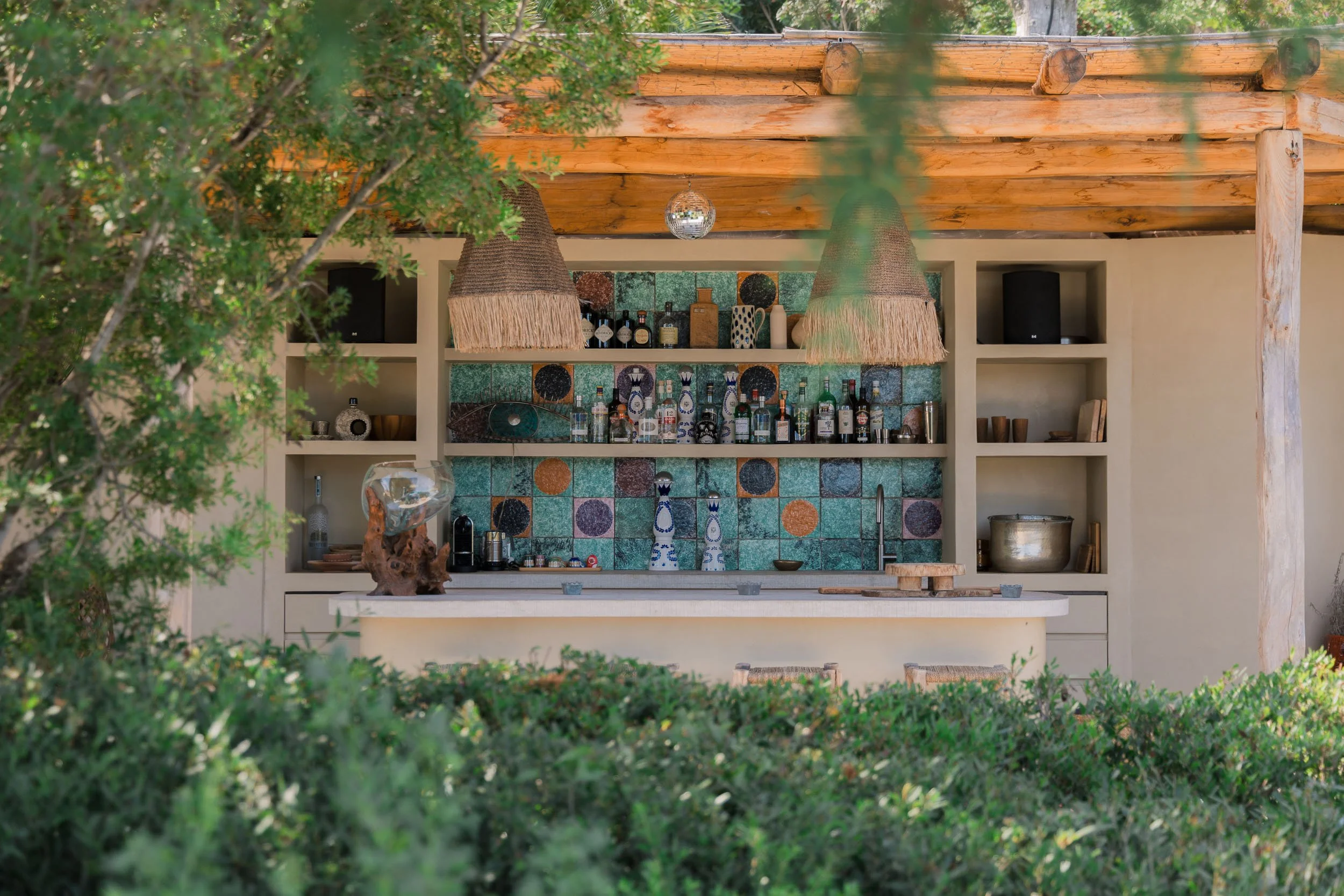 Outdoor bar with colorful tiled backsplash, open shelving with bottles and decor, wooden beam roof, hanging woven light fixtures, and surrounding greenery.