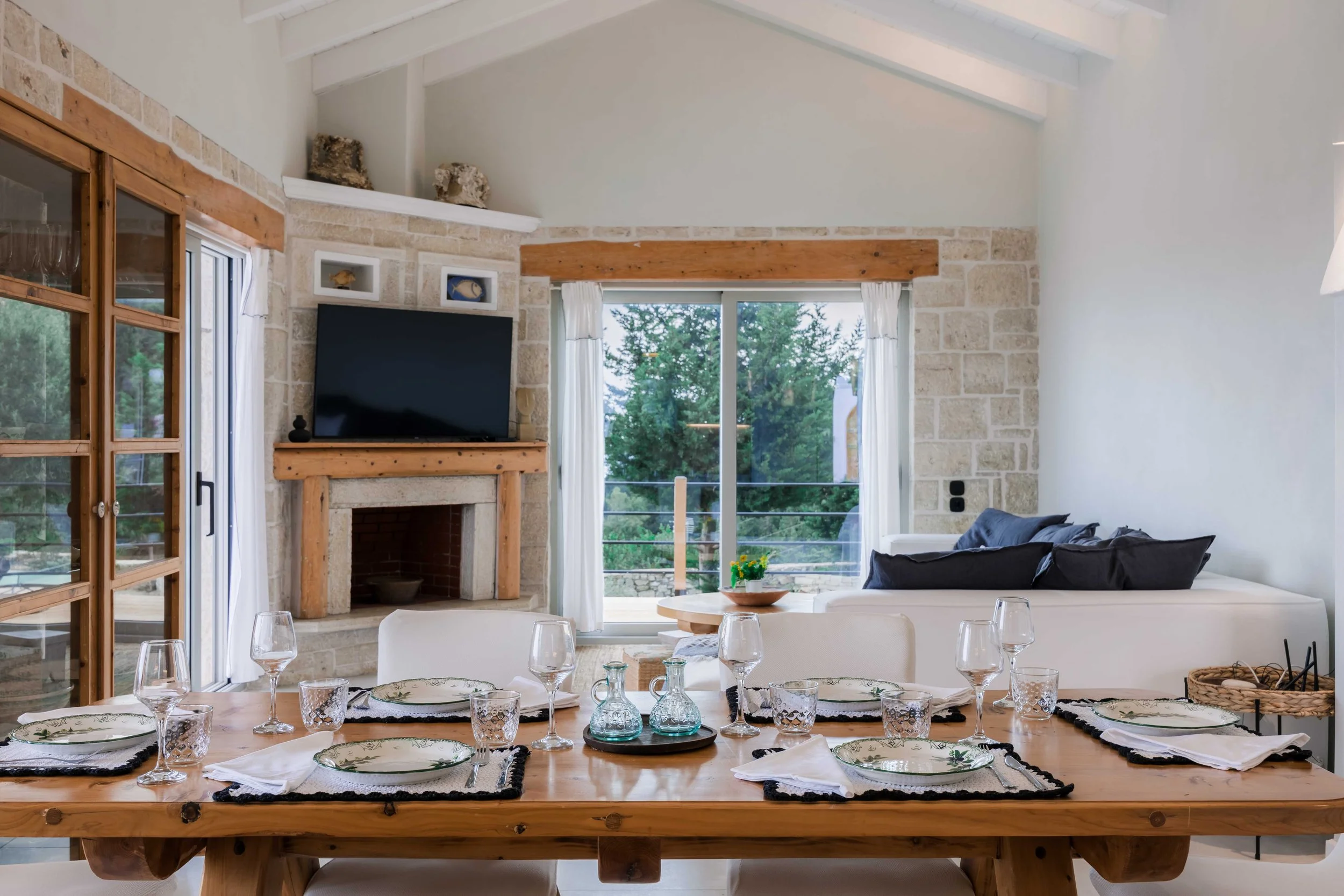 Dining area with a wooden table set with plates, glasses, and napkins, a white couch with dark pillows, a sliding glass door with curtains leading to an outdoor view, a stone fireplace, and a television above the fireplace.