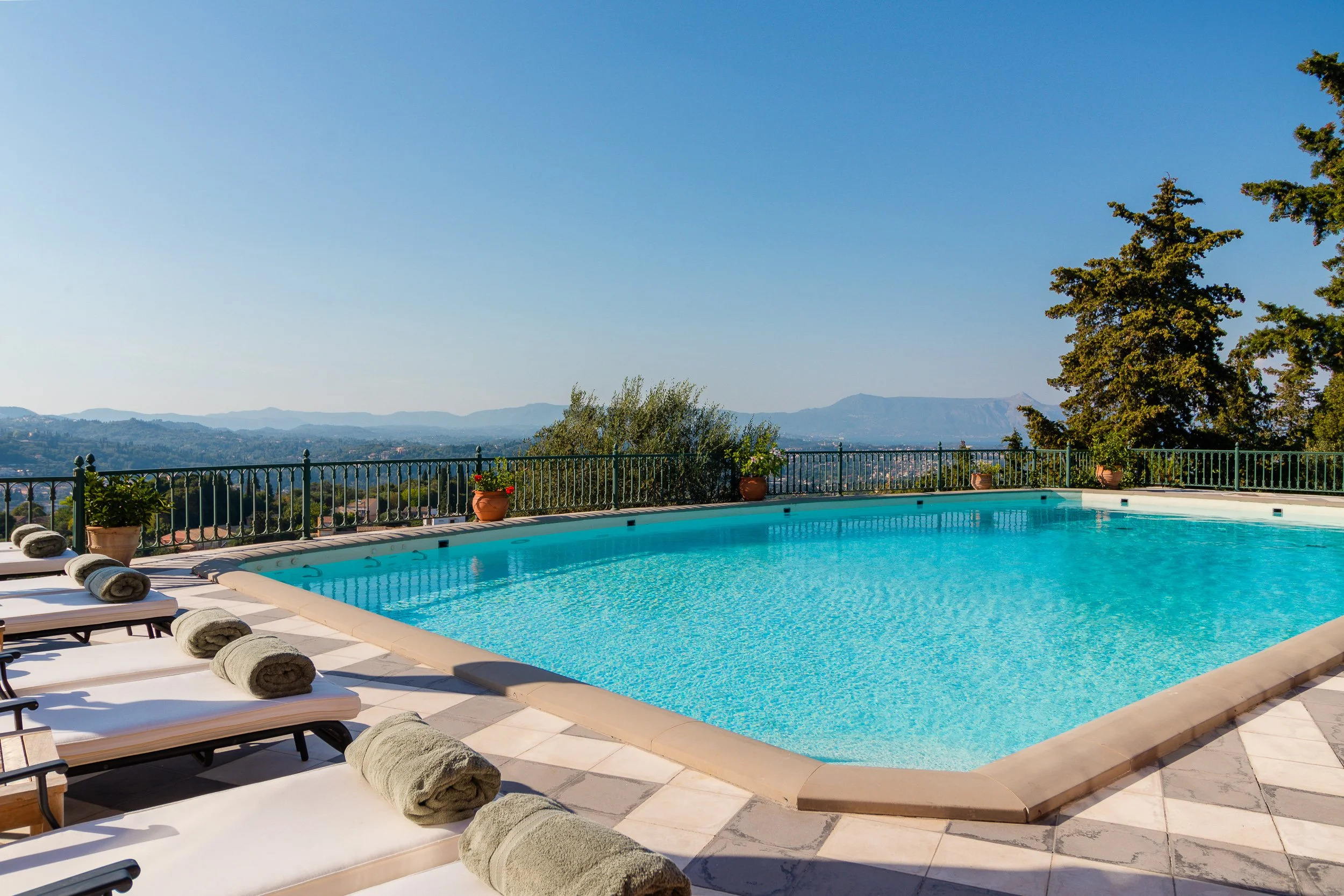 Outdoor swimming pool with lounge chairs and rolled towels, surrounded by potted plants and a metal fence, overlooking a scenic landscape with hills and trees under a clear blue sky.