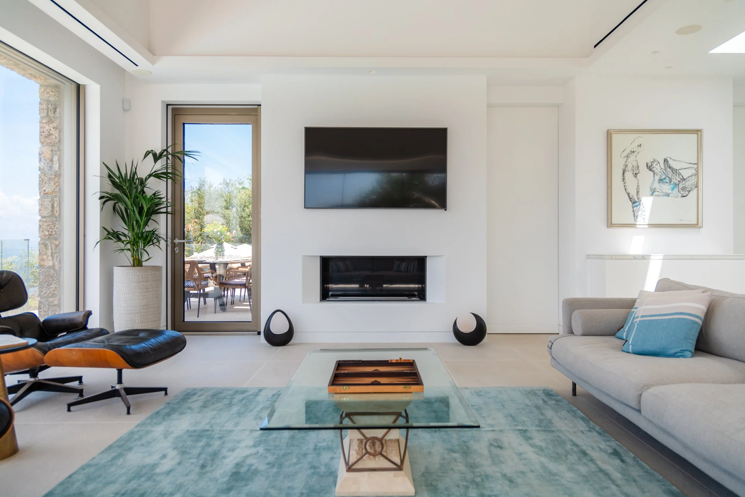 Modern living room with a wall-mounted TV above a fireplace, a glass coffee table with a wooden backgammon board, a gray sofa with a blue striped pillow, black curved speakers, large windows and a door opening to a balcony with outdoor seating and gr