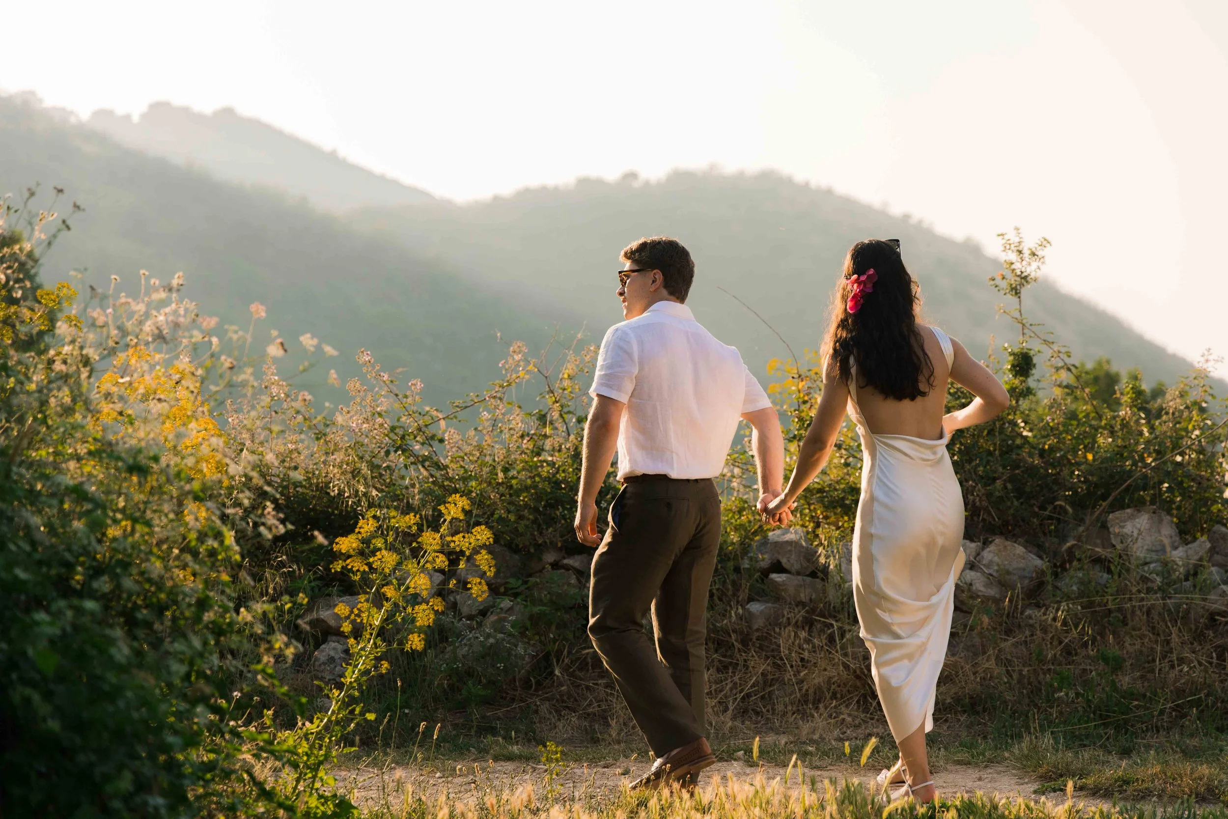 A couple walks hand-in-hand outdoors in a natural setting with mountains and greenery in the background, during sunset.