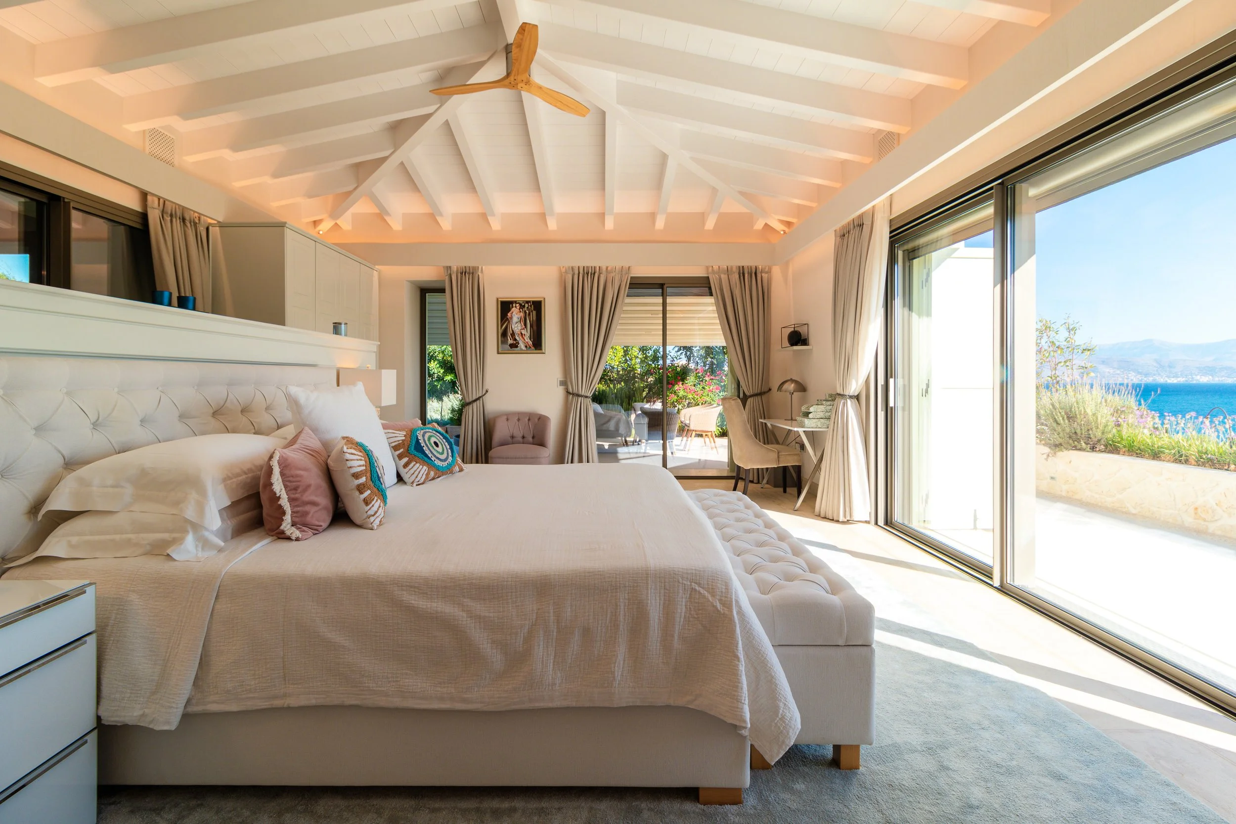 Luxury bedroom with white tufted bed, decorative pillows, sliding glass doors opening to a balcony with outdoor seating, and a view of the sea and mountains.
