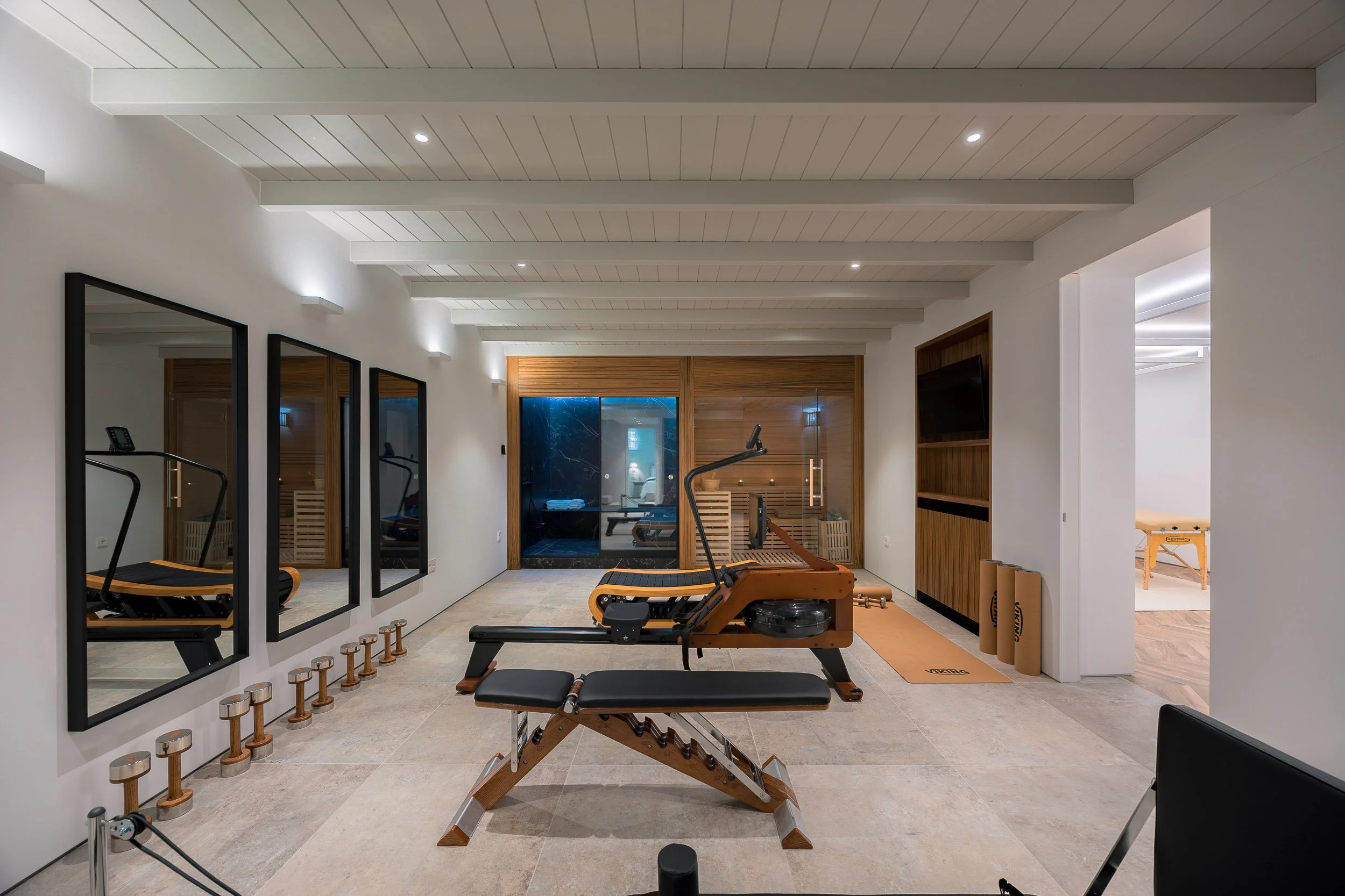 Indoor home gym with exercise equipment including a rowing machine and an adjustable weight bench, mirrors on the left wall, wooden accents, and a sliding glass door at the back leading to a sauna room.