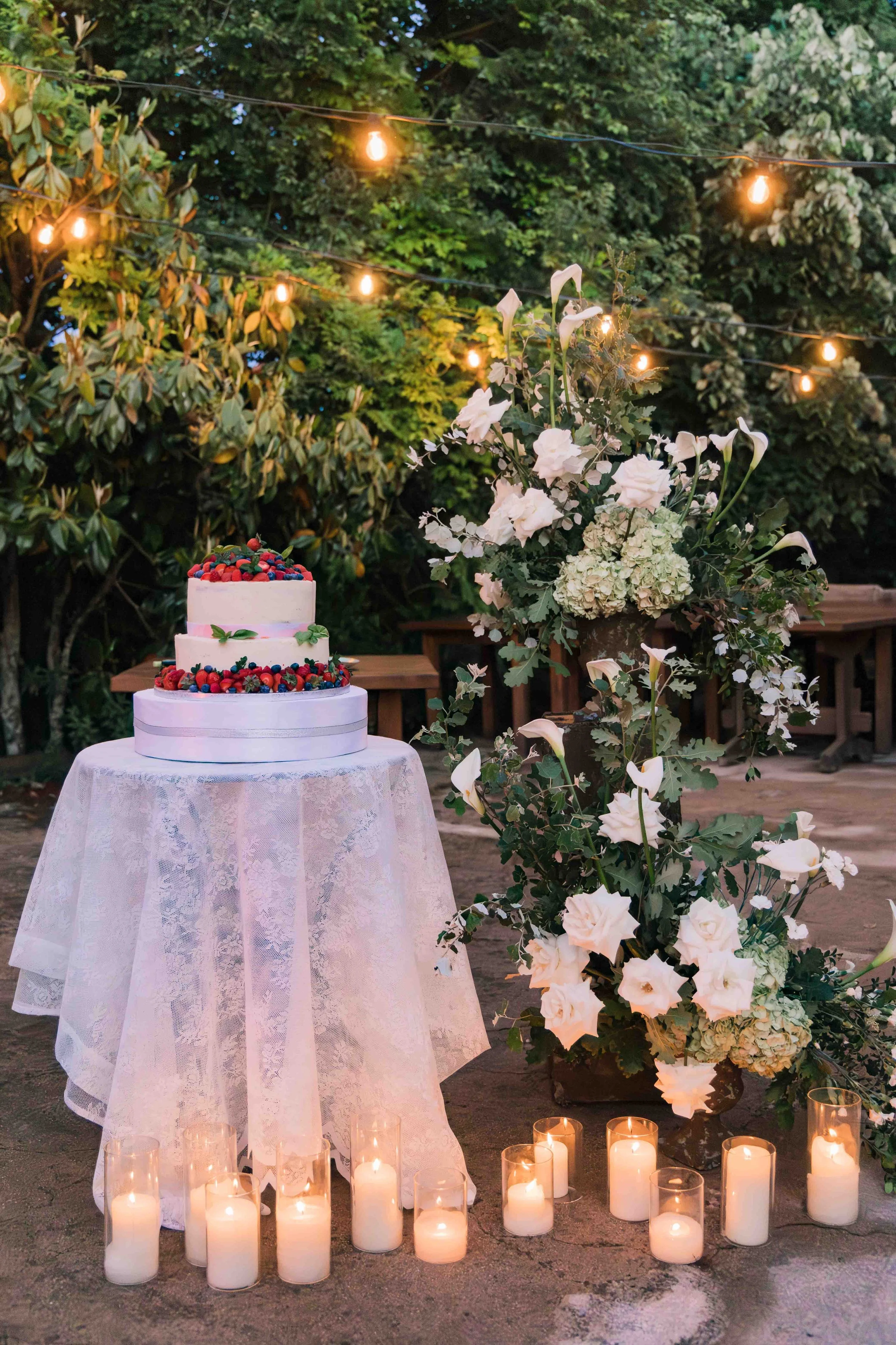 A three-tier white wedding cake decorated with berries on a lace-covered table, surrounded by white candles in glass holders, with a large floral arrangement of white flowers and greenery next to it, set outdoors with string lights and trees in the b