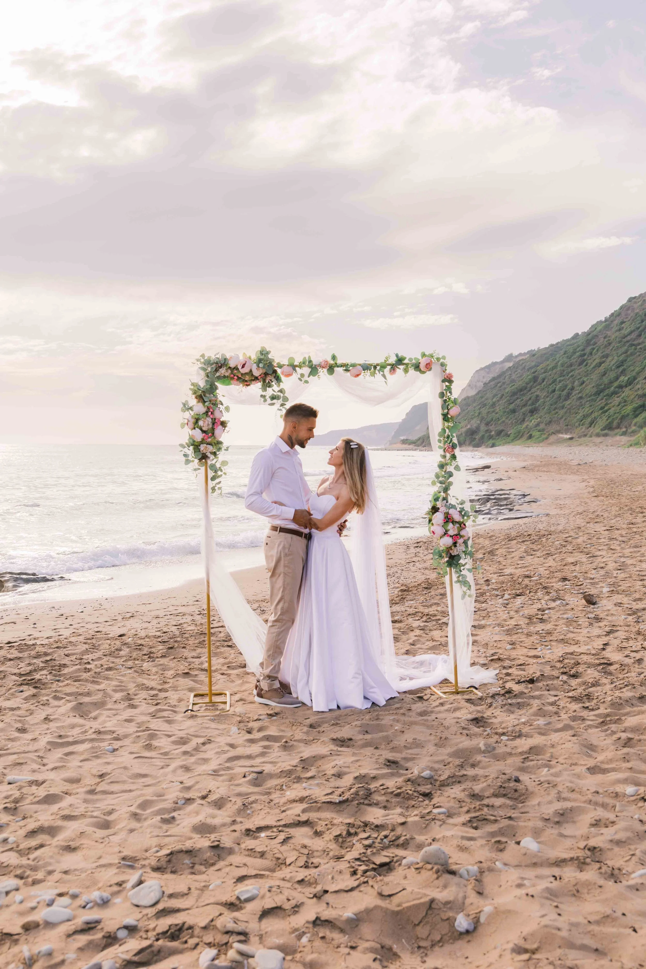 A bride and groom standing on the beach, holding hands and smiling, framed by a floral wedding arch with pink and white flowers and greenery, with a scenic shoreline and hills in the background during sunset.